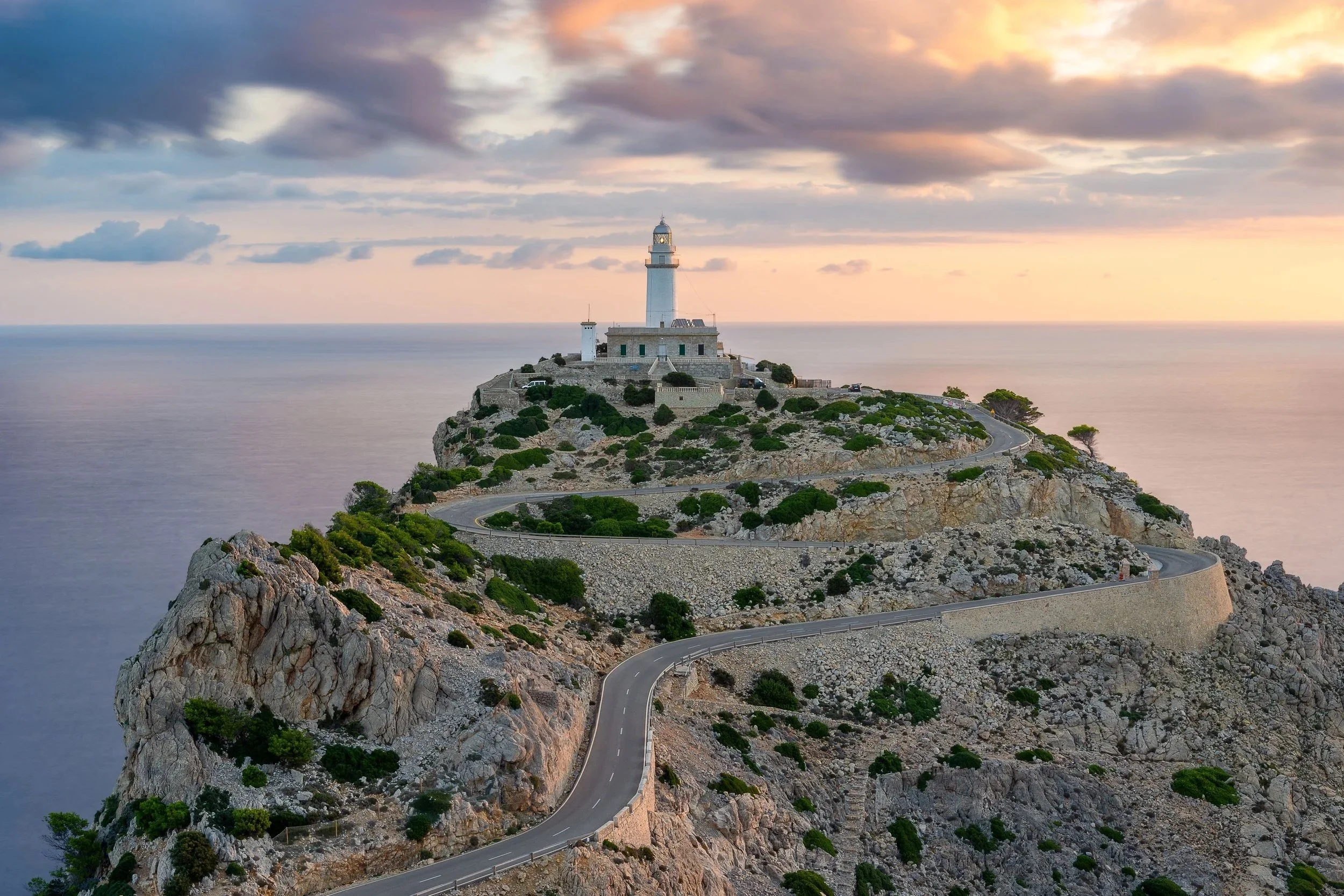 A lighthouse on a rocky hill with a winding road leading up to it, surrounded by the sea, under a colorful sunset sky.