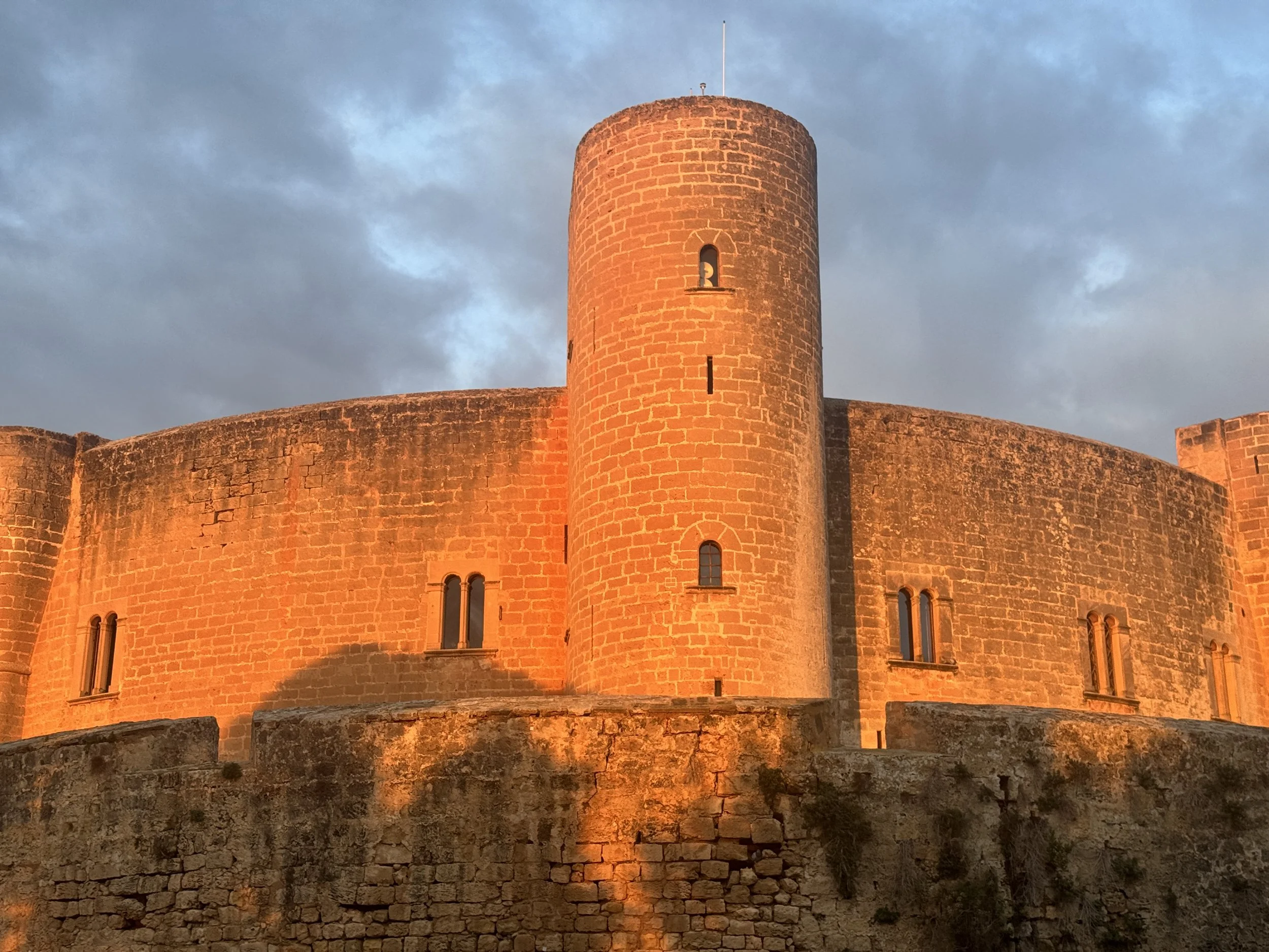 A stone castle with a large round tower, illuminated by the sunset, against a cloudy sky.
