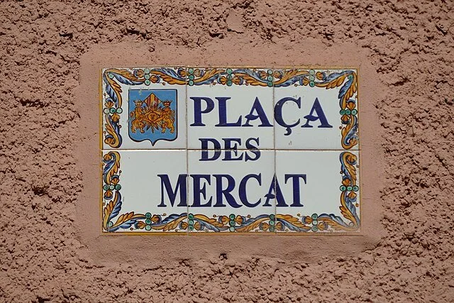 Decorative ceramic street sign reading "Plaça des Mercat" on a textured pink wall.