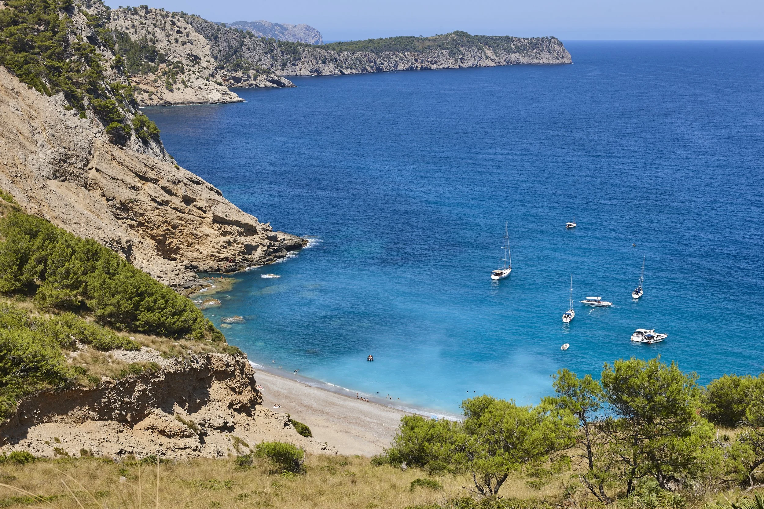 A scenic coastal view of a bay with turquoise water, anchored sailboats, a curved beach, rocky cliffs, and green vegetation on the hillside.