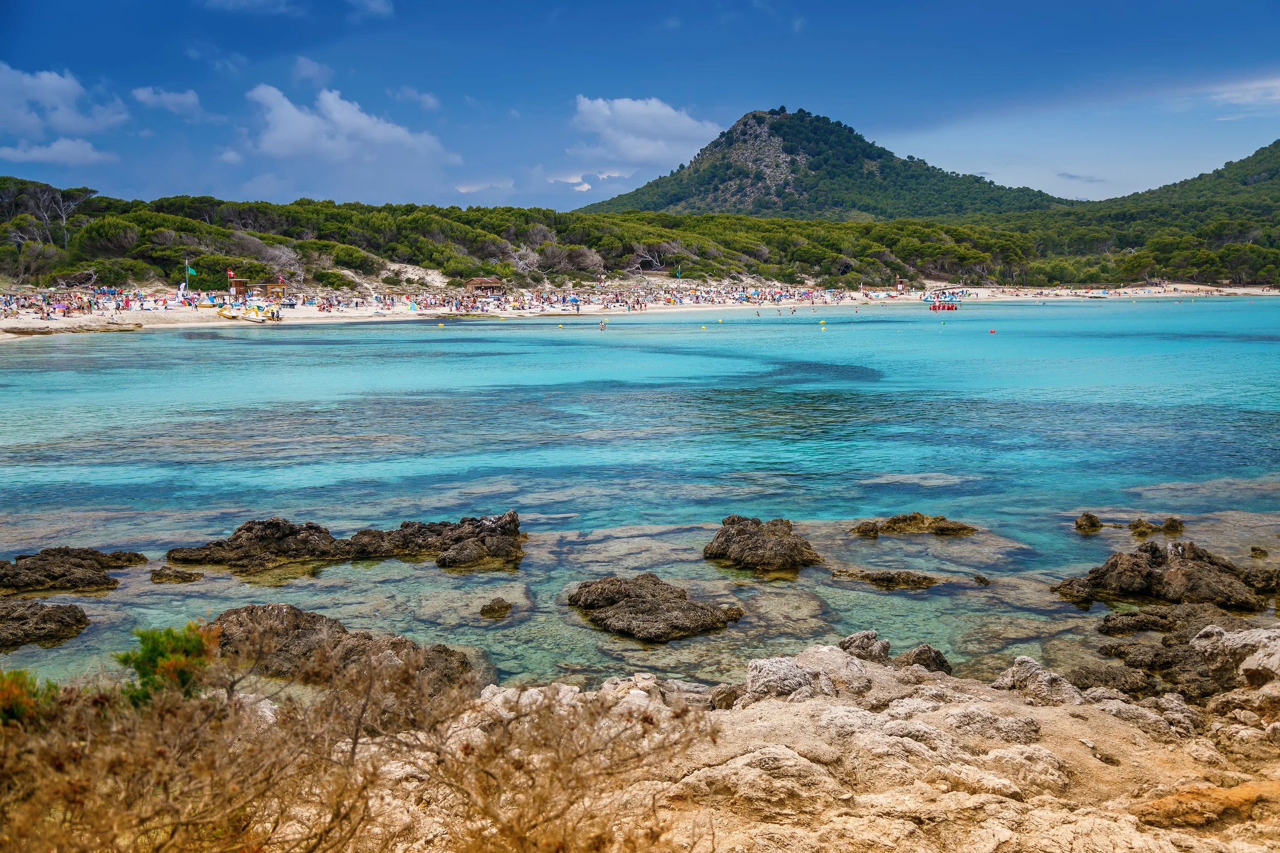 Scenic view of a beach with turquoise water, rocky shoreline in the foreground, sandy beach with sunbathers and umbrellas, green forested hills and a mountain in the background under a partly cloudy sky.