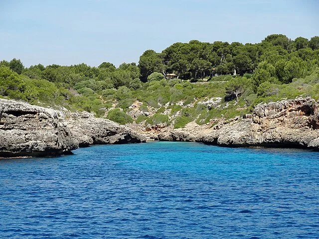 A blue waterway with rocky cliffs on both sides and green trees on a hillside in the background under a clear sky.