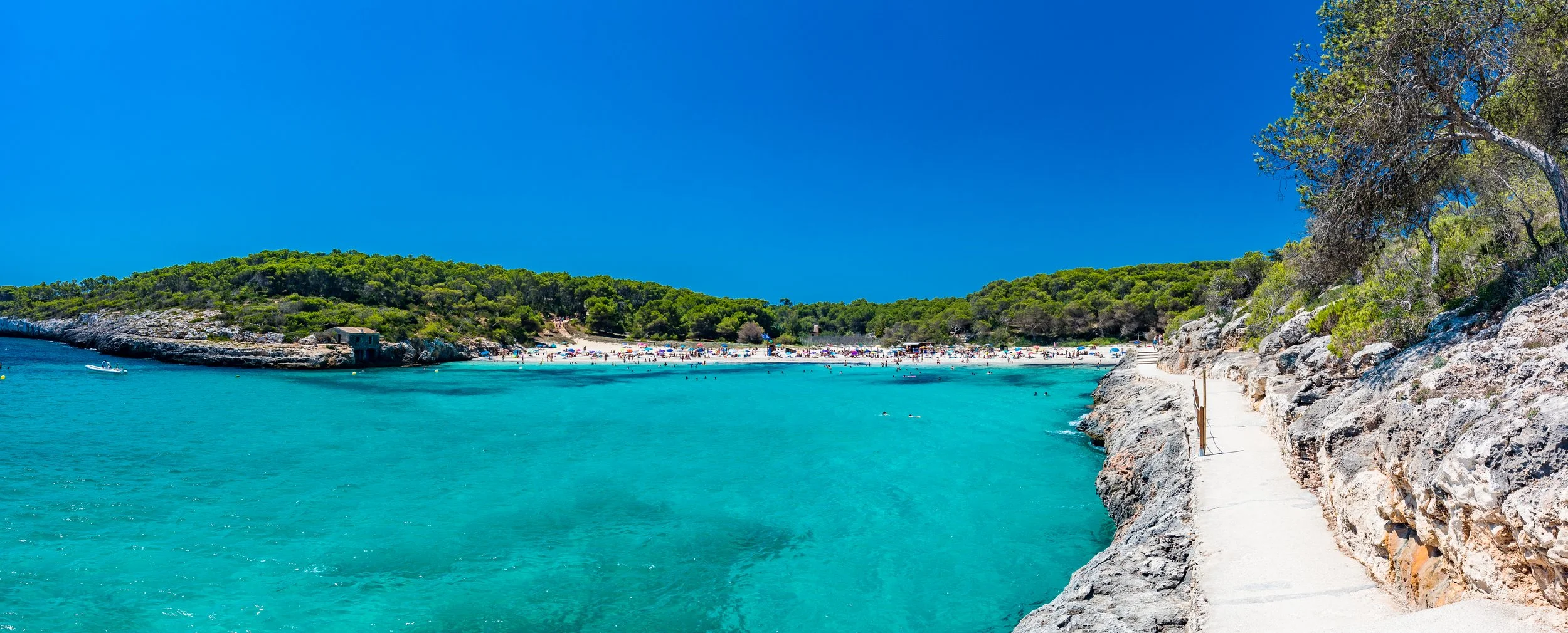 Scenic view of a sandy beach with colorful umbrellas and people, turquoise water, rocky cliffside with greenery, and a clear blue sky.