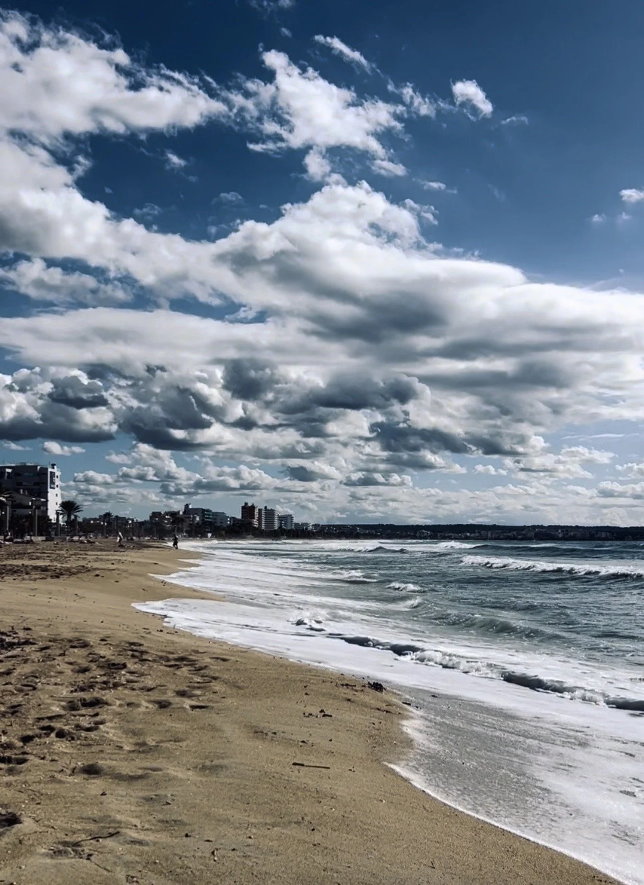 A sandy beach along the ocean with waves crashing onto the shore, under a partly cloudy sky with buildings visible in the distance.