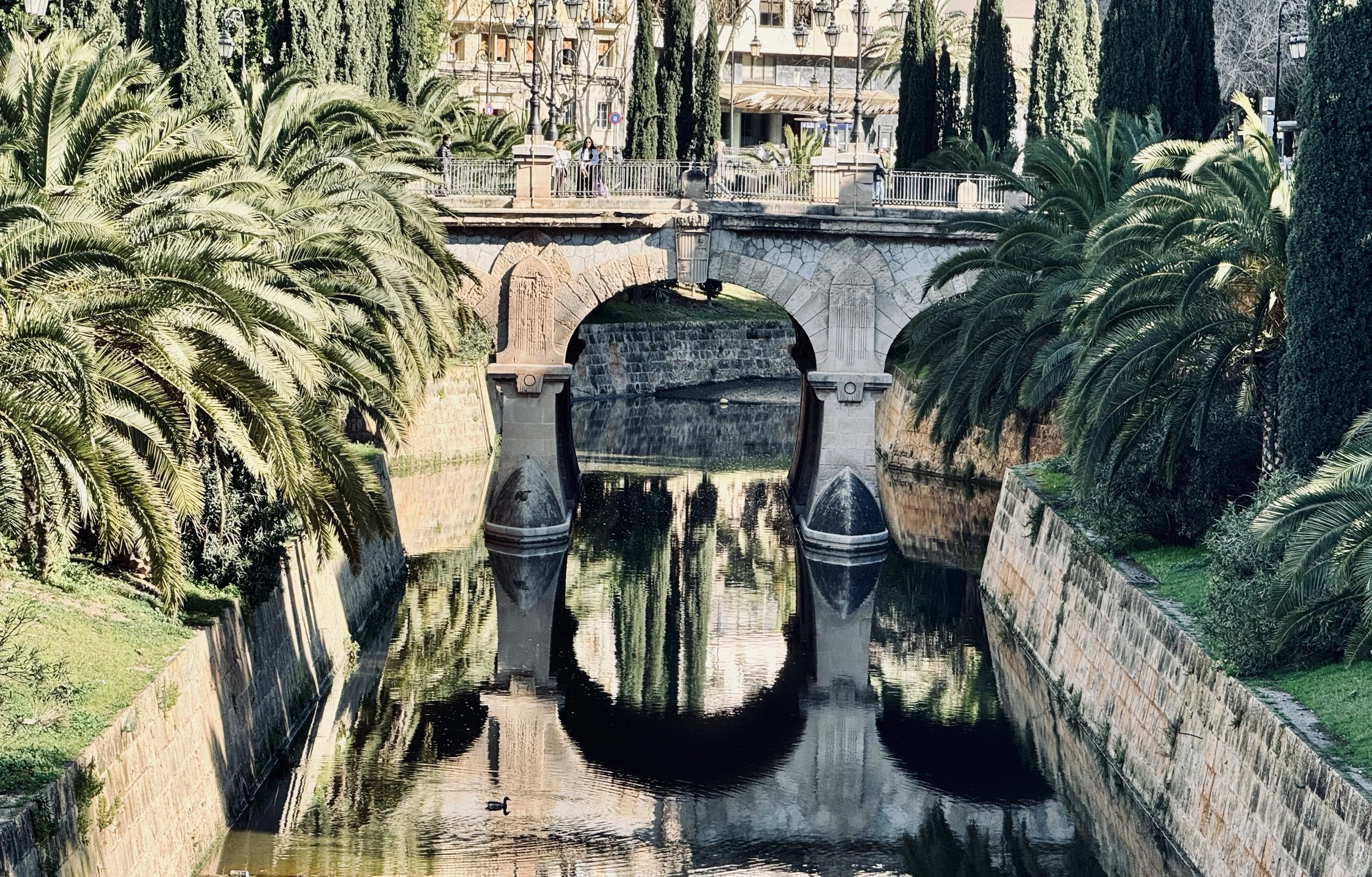 A stone arched bridge over a canal, lined with lush green palm trees and tall cypress trees. Pedestrians walk on the bridge, and reflections of the bridge and trees can be seen in the water below.