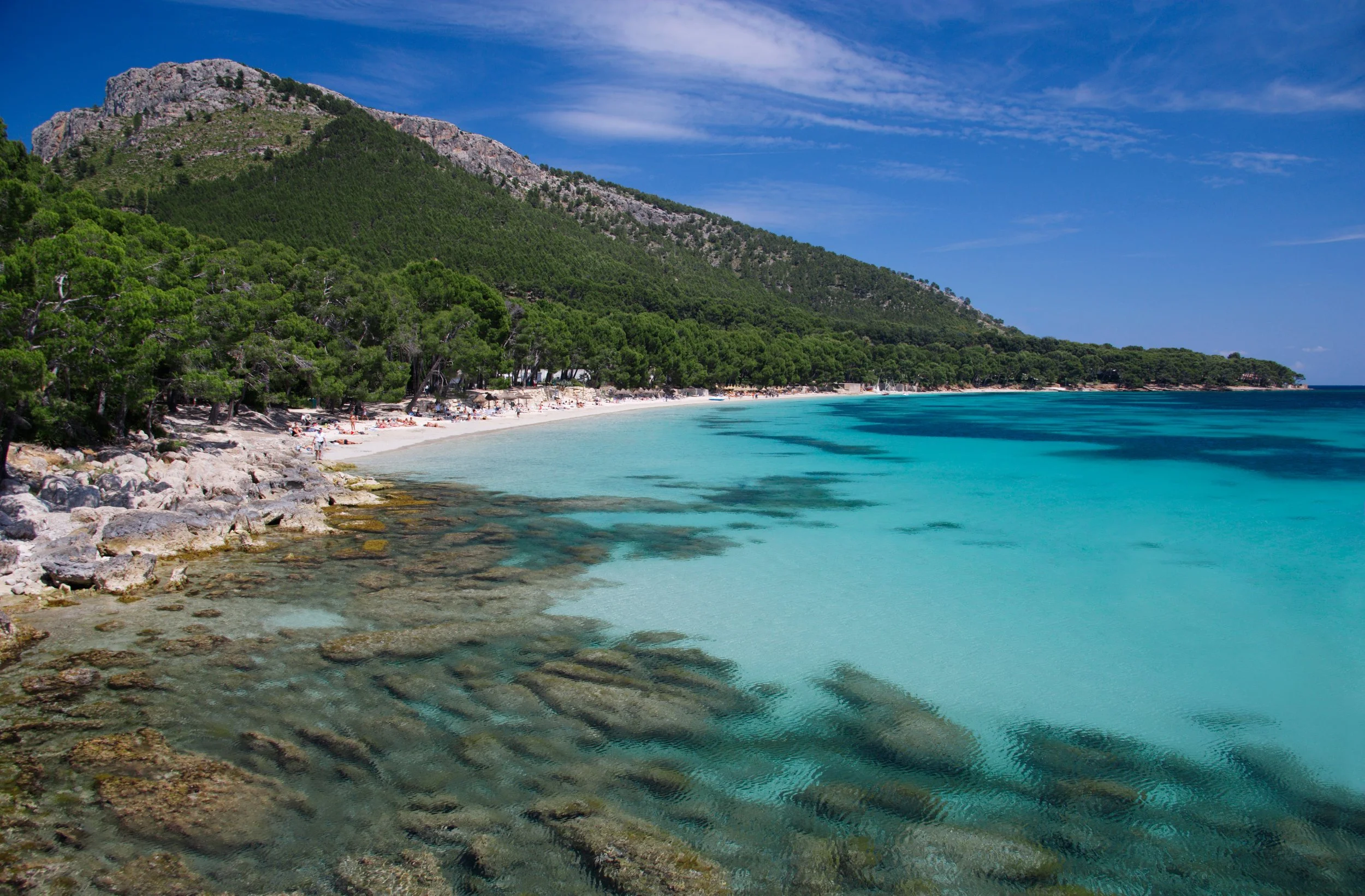 Sunny beach with turquoise water, rocky shoreline, green forested hill, and blue sky with scattered clouds.