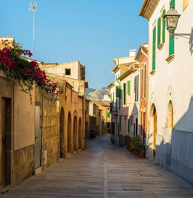 A quiet narrow street in a Mediterranean town with colorful buildings, green shutters, flower pots, and blue sky