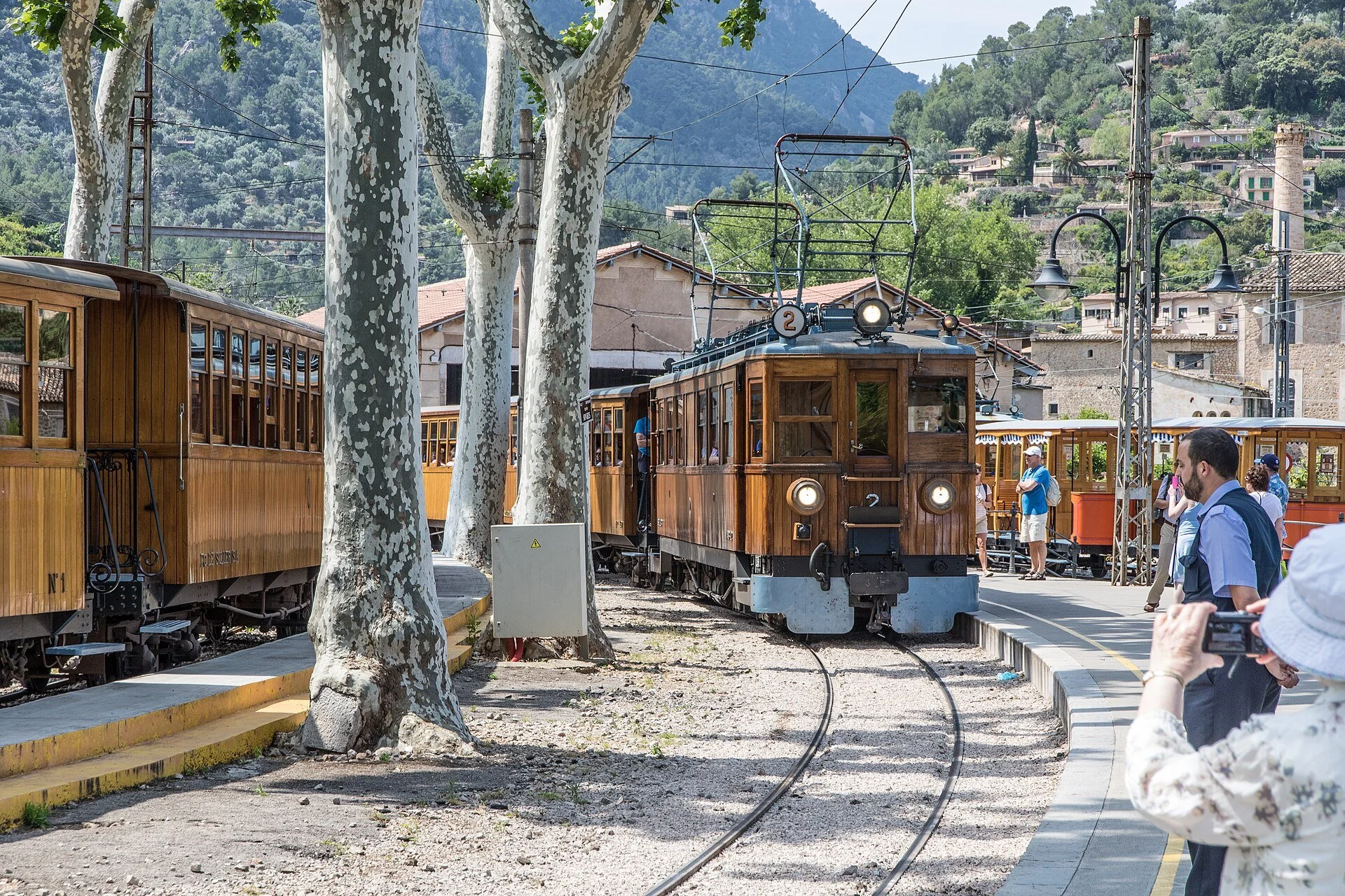 A vintage wooden train arriving at a station with onlookers taking photos, surrounded by trees and hilly terrain in the background.