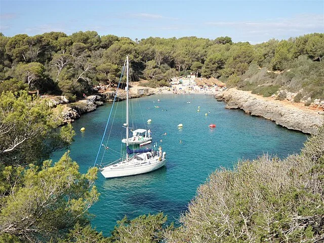 A small sailboat anchored in a clear, calm cove with rocky shores and lush green trees surrounding the water, dotted with buoys.