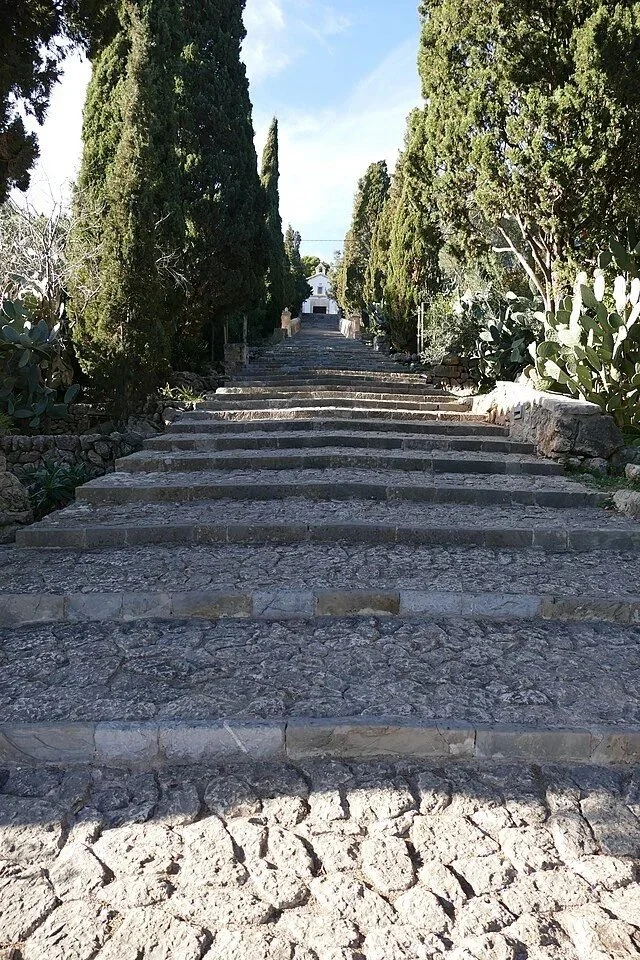 Stone steps ascending through a garden of tall, green cypress trees and cactus, leading towards a white building.