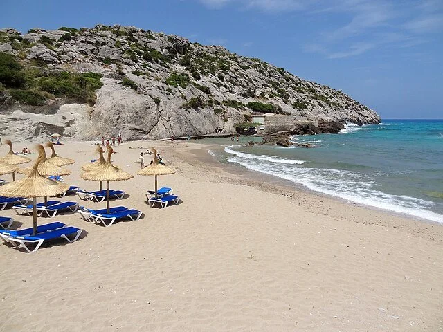 Sandy beach with blue lounge chairs and straw umbrellas, rocky hillside and people in the background, turquoise sea, clear blue sky.