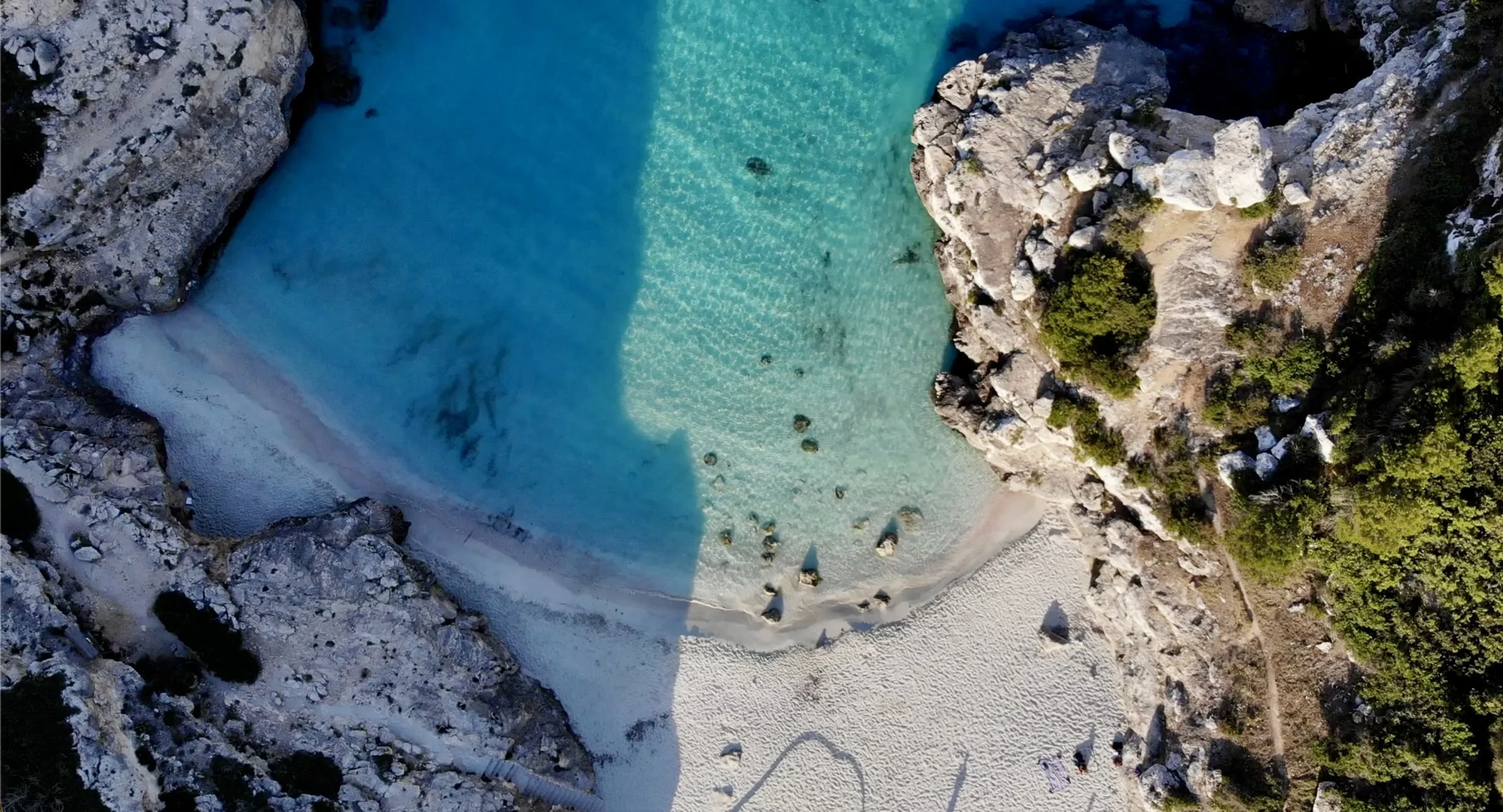 An aerial view of a secluded blue lagoon surrounded by rocky cliffs and green vegetation with a sandy beach.