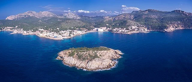 Aerial view of an island off the coast with a town along the shoreline, mountains in the background, and clear blue water.