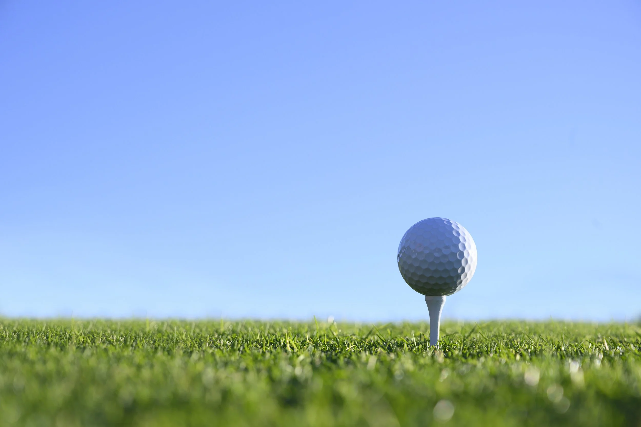 A close-up of a golf ball on a tee, set on a grassy golf course under a clear blue sky.