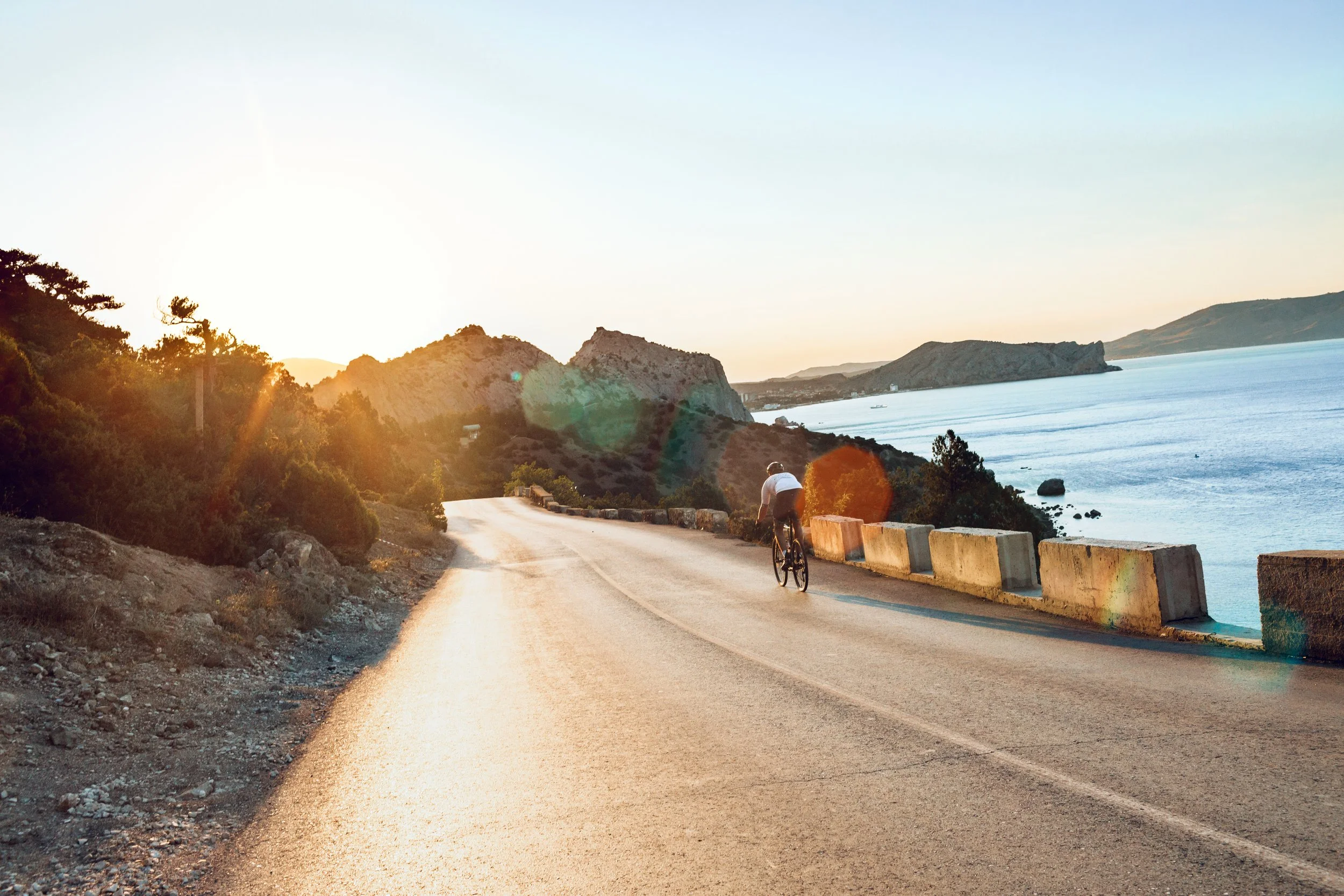 A person riding a bicycle on a coastal road during sunset, with water and hills in the background.
