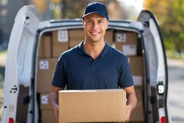 A man in a blue shirt and cap holding a cardboard box, standing in front of an open delivery van filled with boxes, outdoors during daylight.