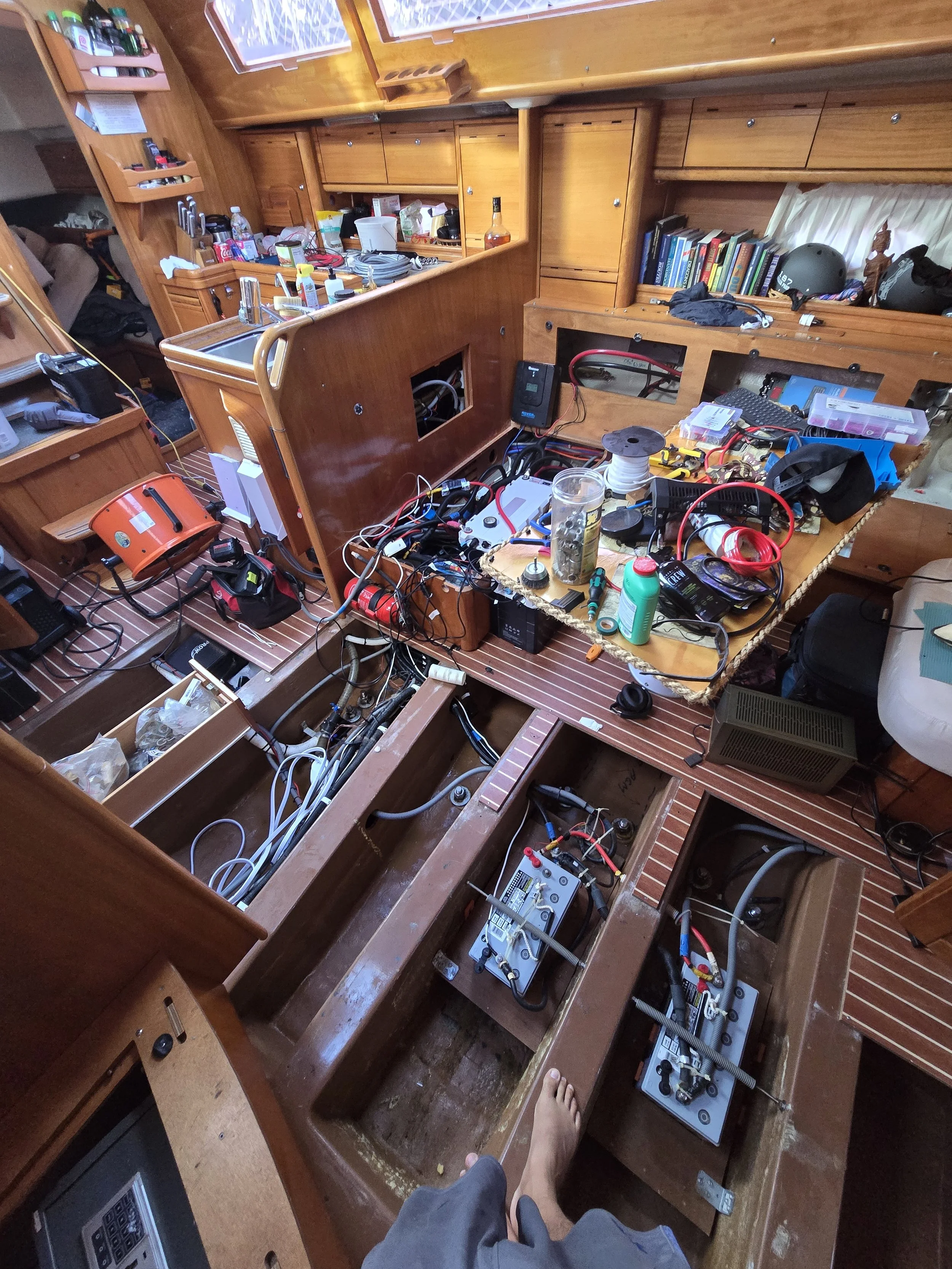 The image shows the interior of a boat with parts of the paneling removed, revealing wiring, electrical components, and empty spaces where appliances or fittings might be installed. There are various tools, wires, and electronic devices scattered across the floor and work surfaces, indicating ongoing maintenance or renovation.