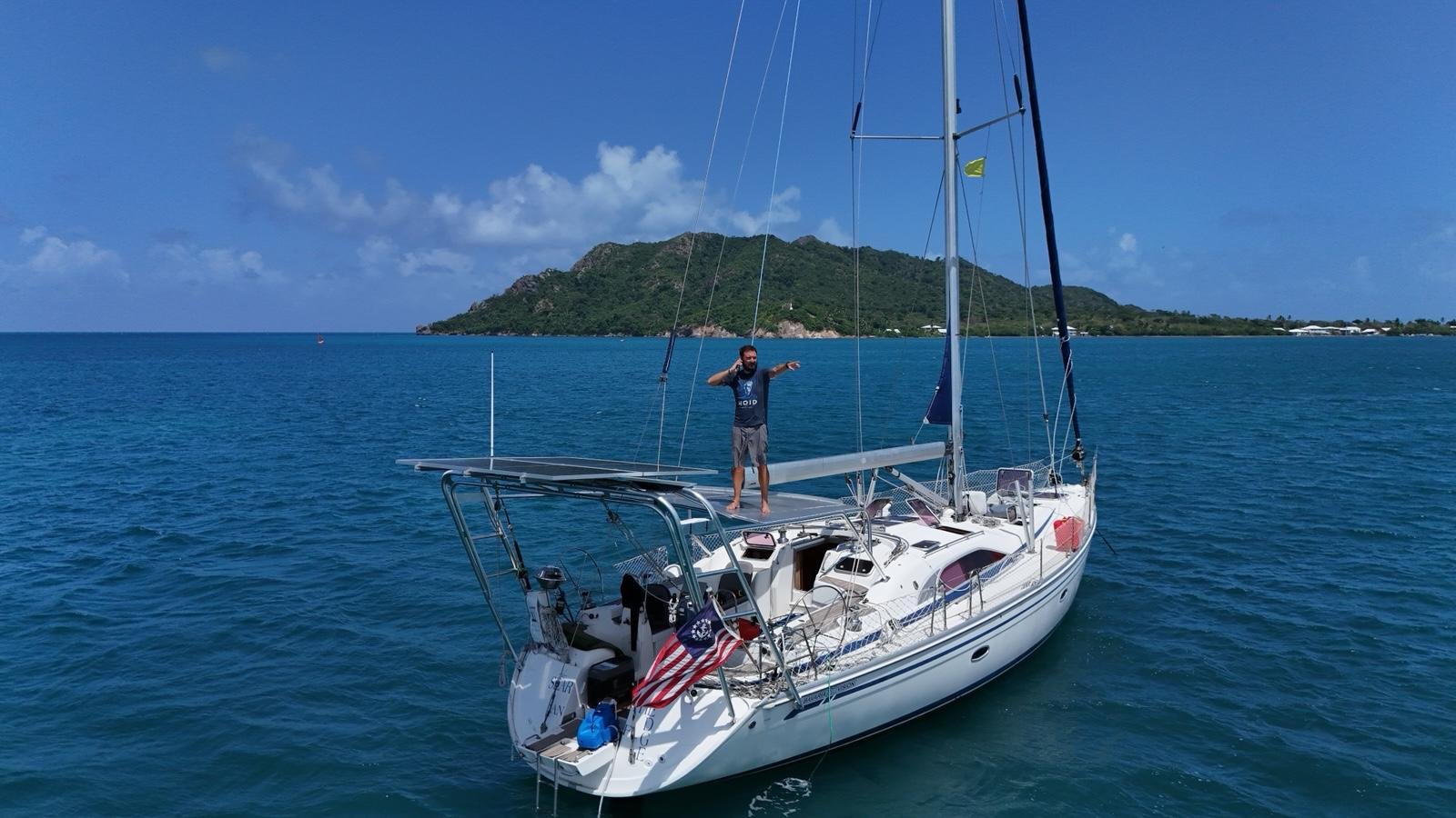 Man standing on the deck of a sailboat in calm blue ocean water, with a green island in the background under a partly cloudy sky.