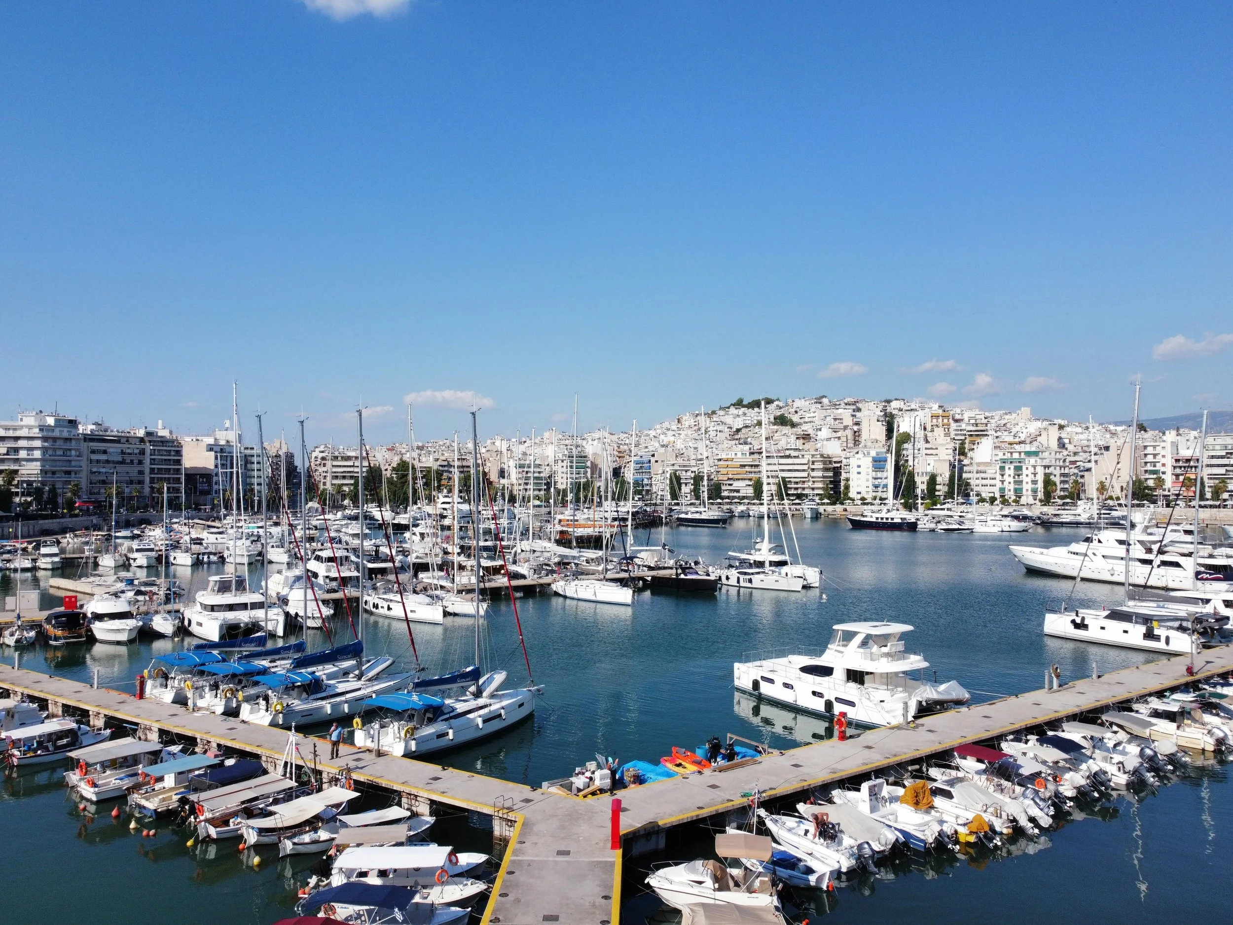 A marina filled with various boats and yachts docked along a pier, with a cityscape of white buildings in the background under a clear blue sky.