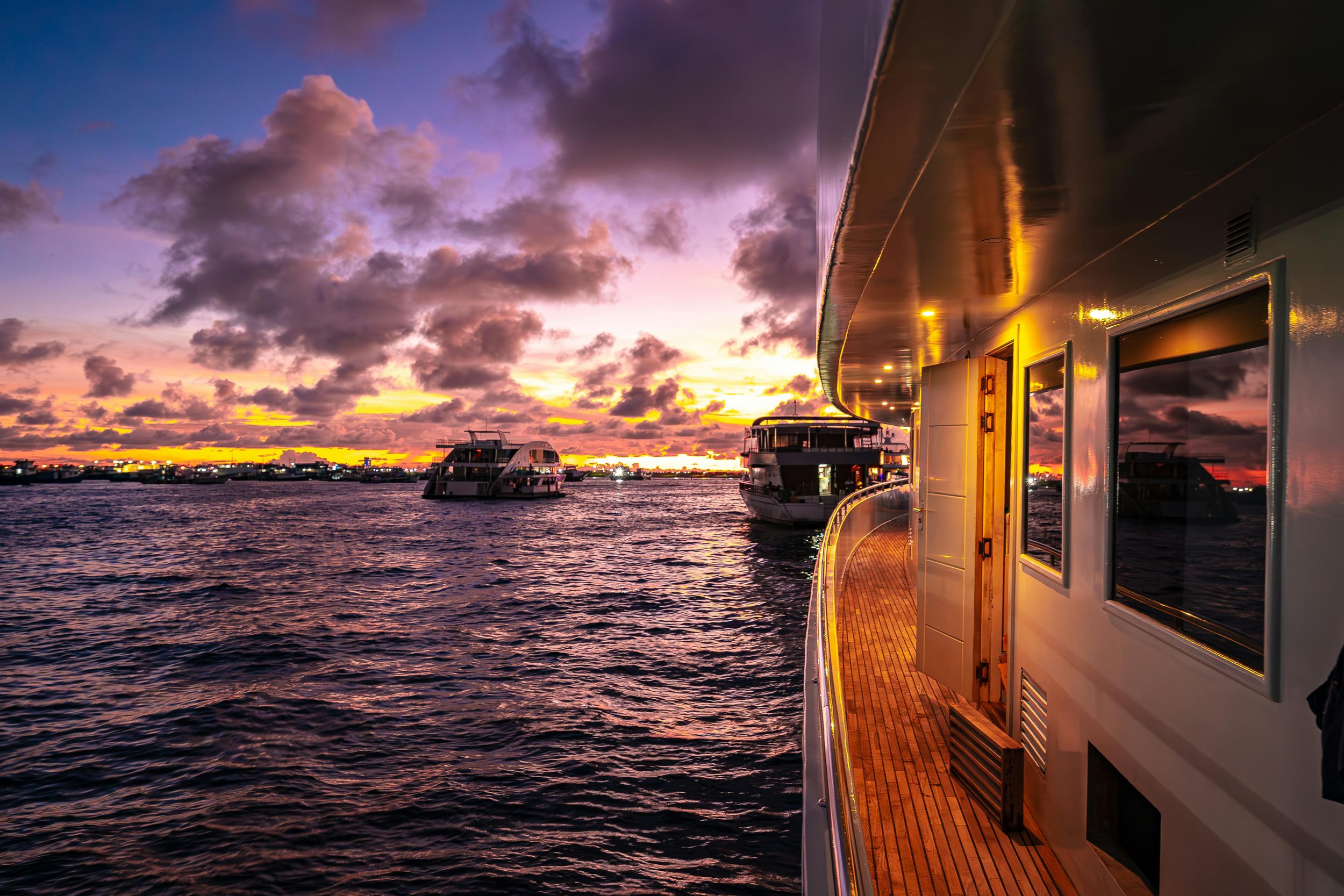 View from a yacht's deck at sunset with other boats on the water and a colorful sky with clouds.
