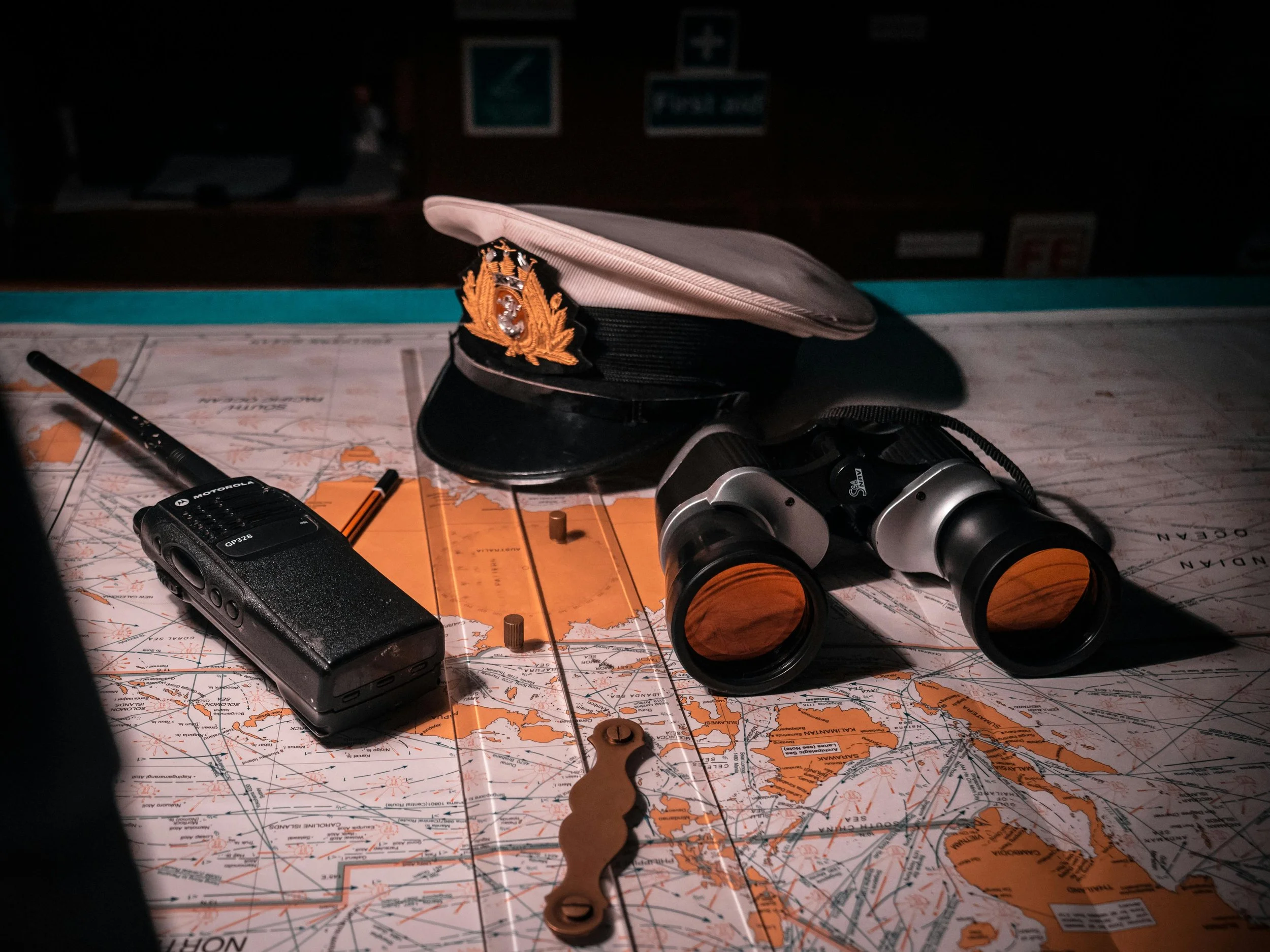 A nautical navigation map spread out on a surface with various items placed on top. These include a英国海军士官的帽子, a small Motorola radio, a pair of binoculars with orange-tinted lenses, a Parker pen, a small metallic tool, and some pins or pegs. The background is dark and out of focus.