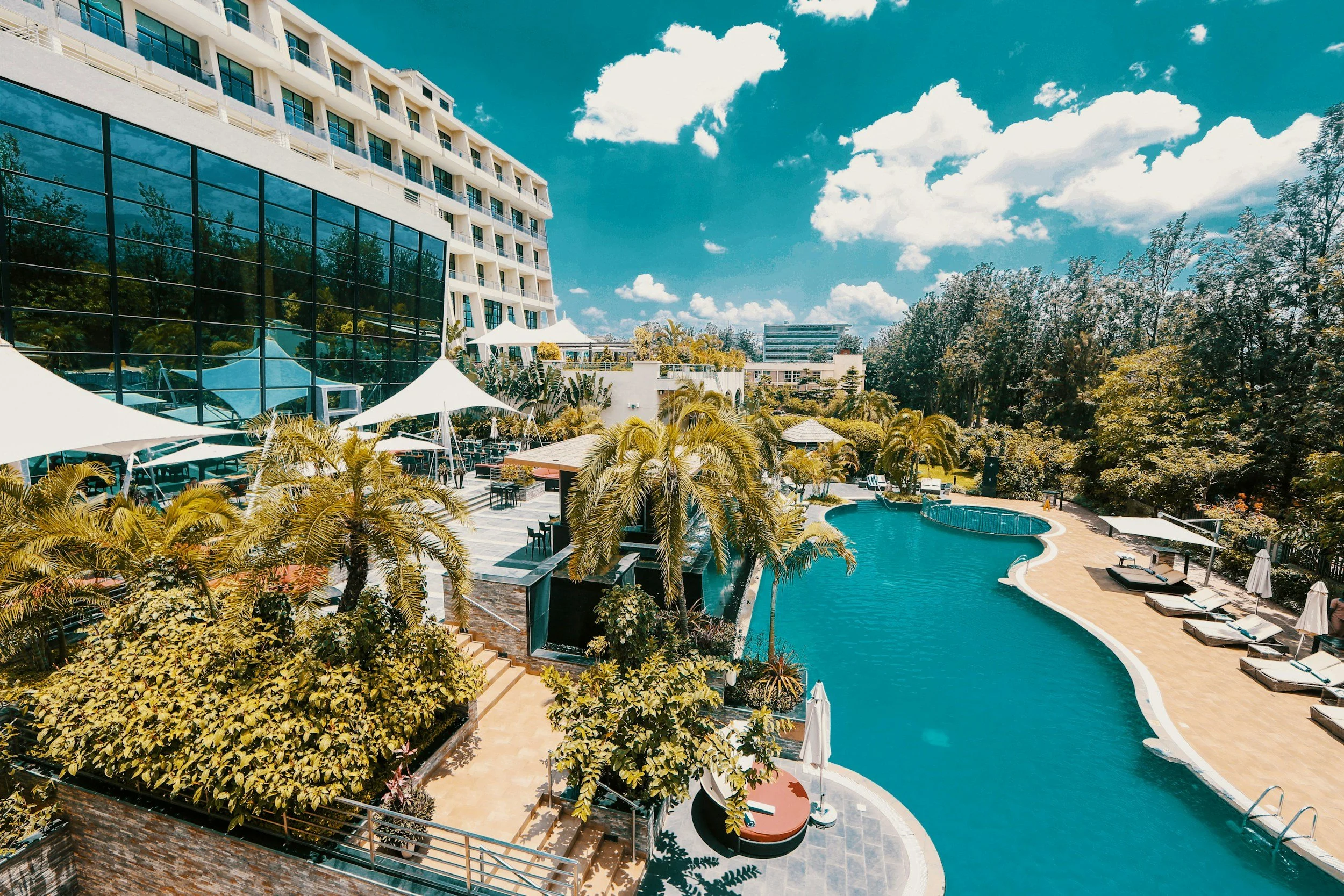 An outdoor swimming pool area at a hotel or resort, surrounded by lounge chairs and umbrellas, with a large modern building and trees in the background under a partly cloudy sky.
