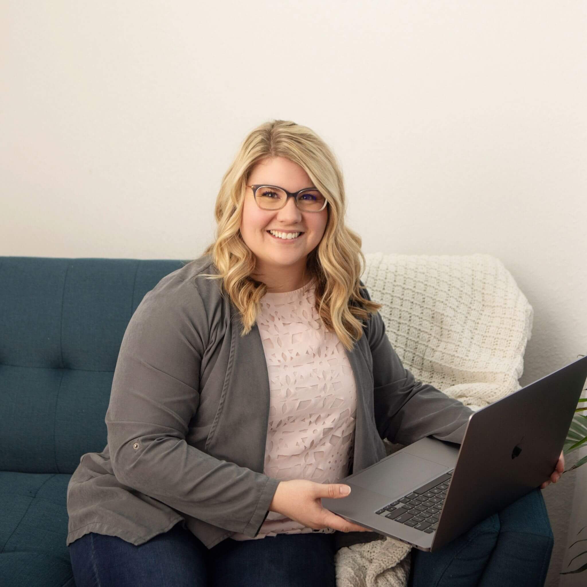 A smiling woman with blonde wavy hair, glasses, sitting on a teal couch, holding a laptop, with a white blanket on the couch behind her.