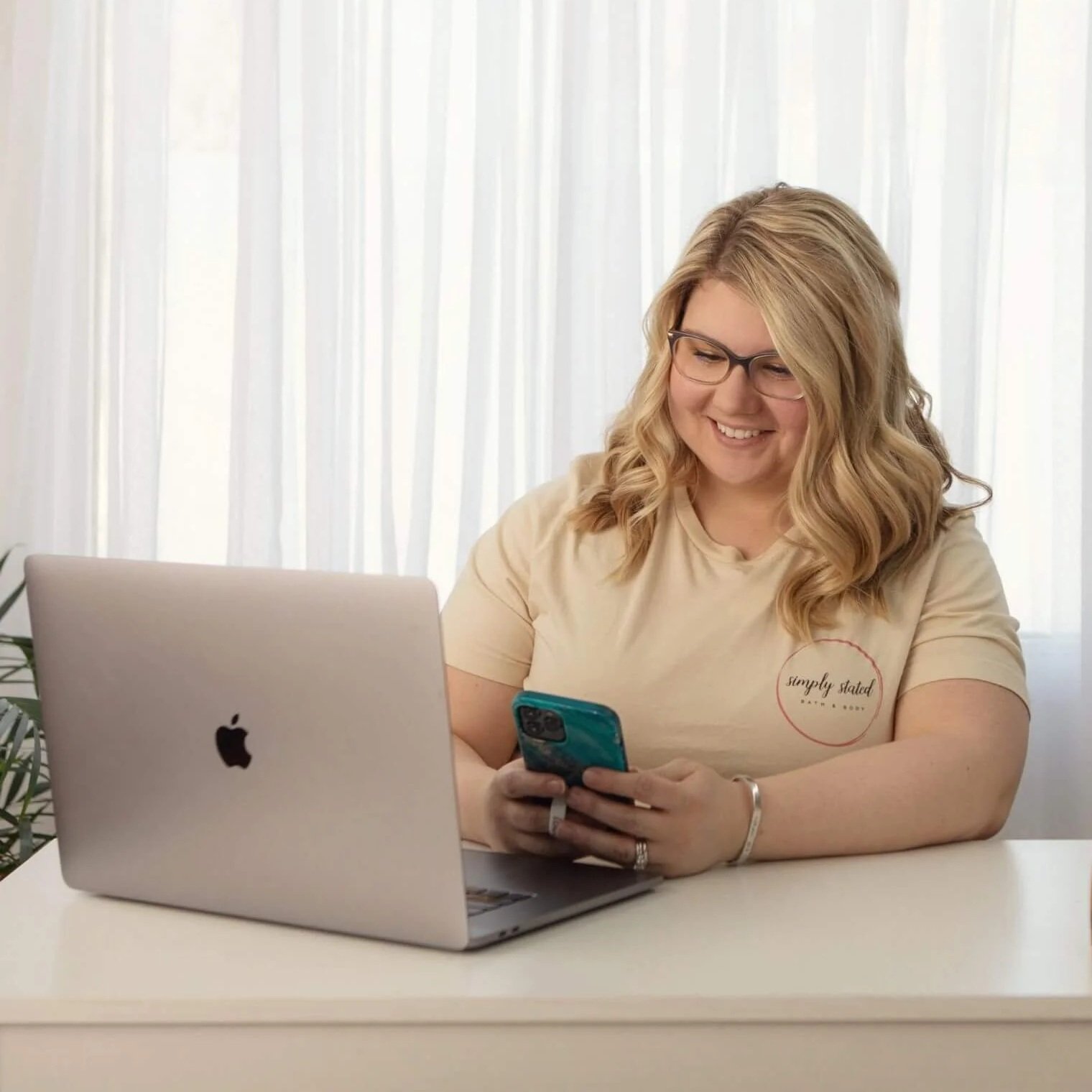 A woman with blonde curly hair and glasses sitting at a white table, looking at her smartphone with a smile, with an open silver MacBook in front of her, in a bright room with white curtains.