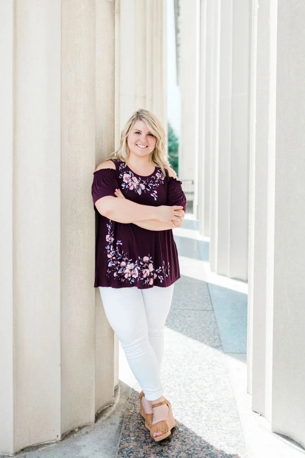 A woman with blonde hair wearing a purple floral blouse, white pants, and tan wedge sandals, leaning against a beige wall outdoors.