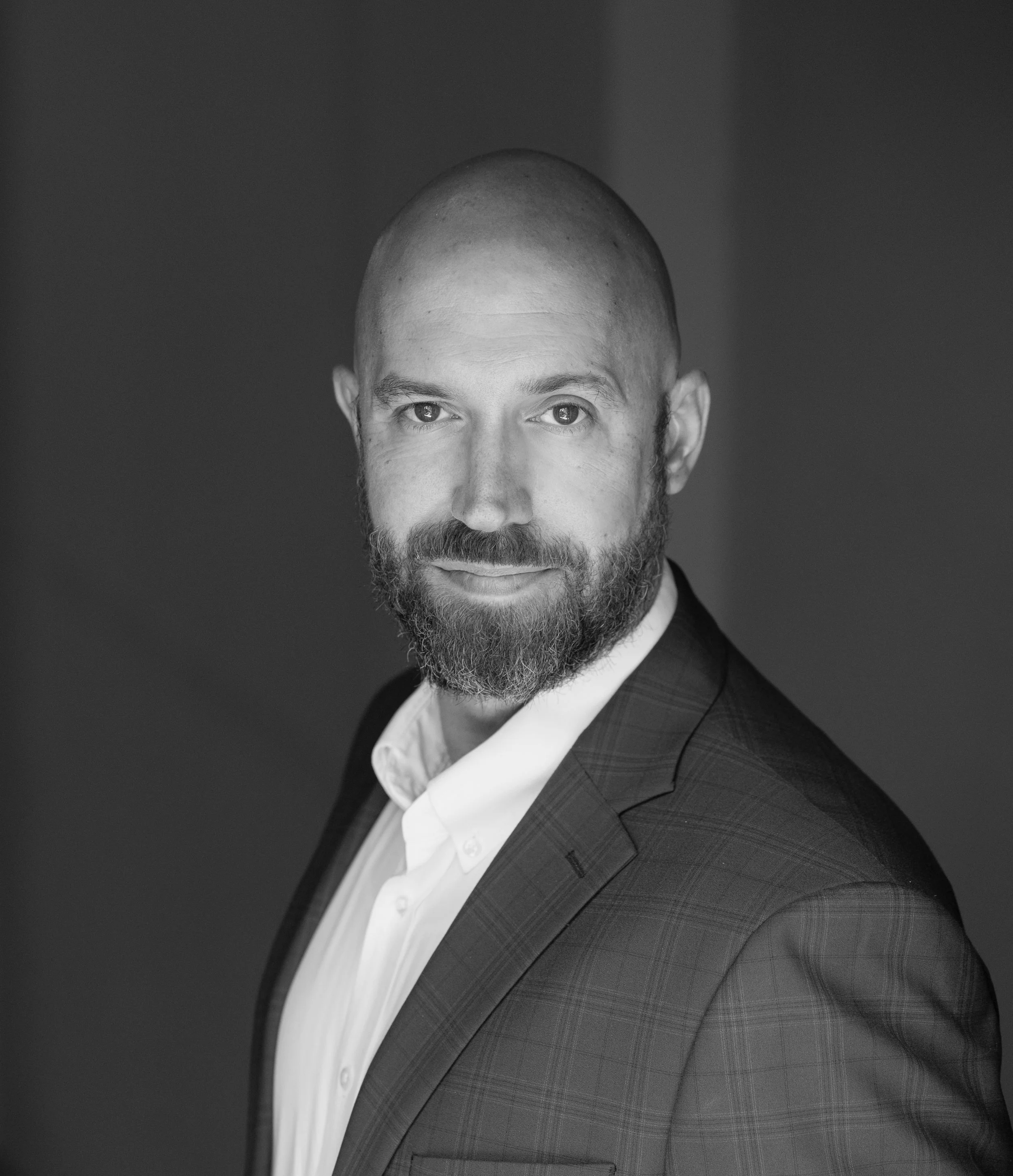 Black and white portrait of a man with a bald head and a beard, wearing a suit jacket and a collared shirt, looking directly at the camera.