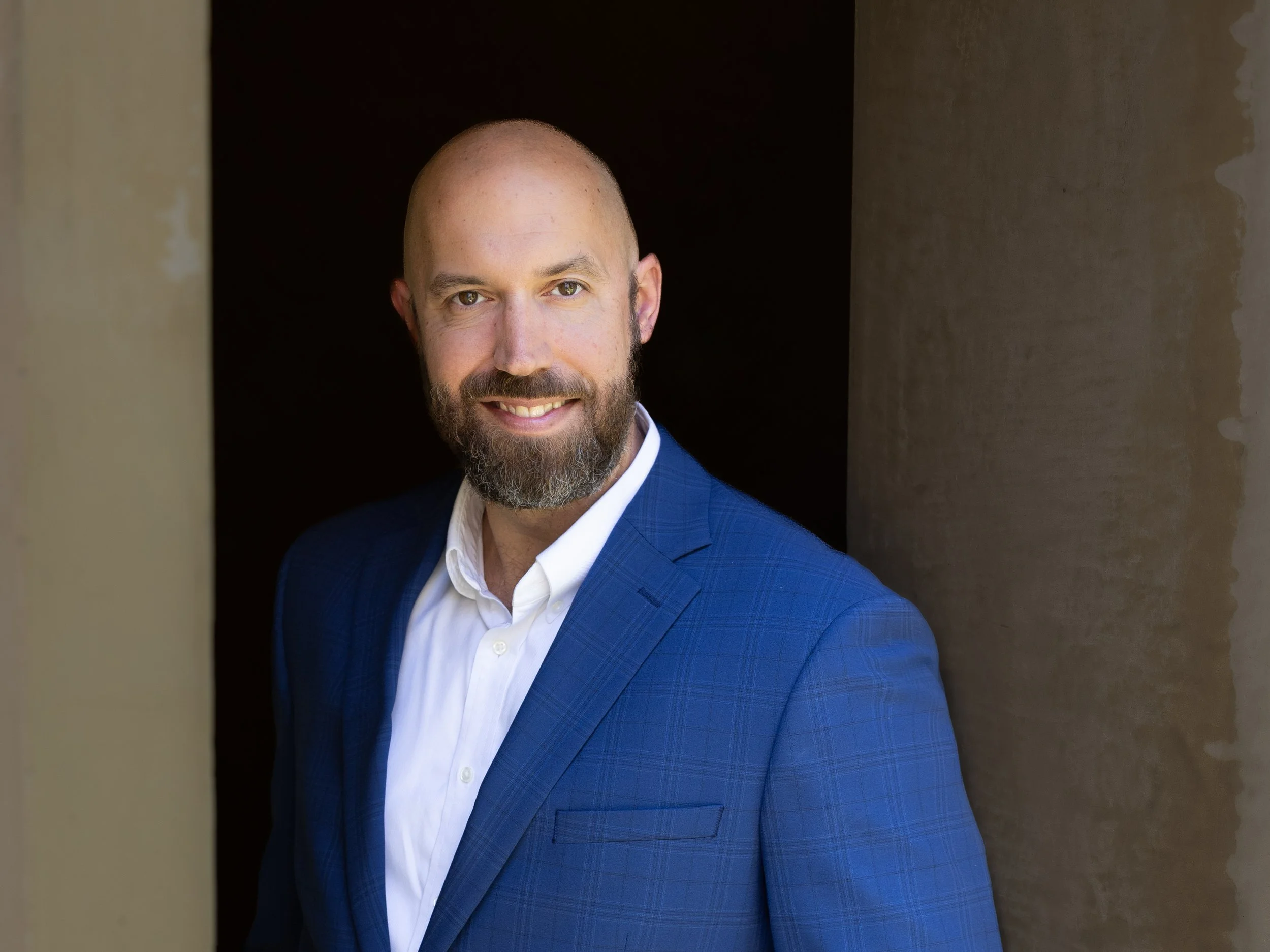 Portrait of a smiling man with a beard in a blue suit and white shirt, standing between two beige columns.