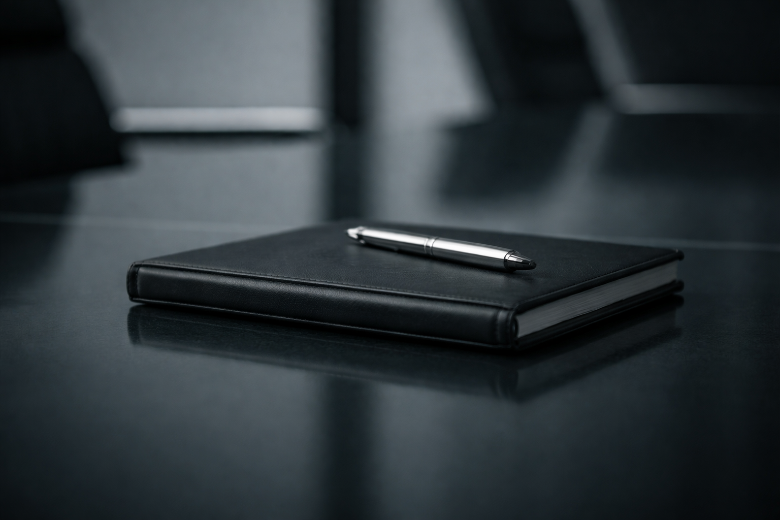 Black notebook with a silver pen on top, placed on a dark table in a dimly lit environment.