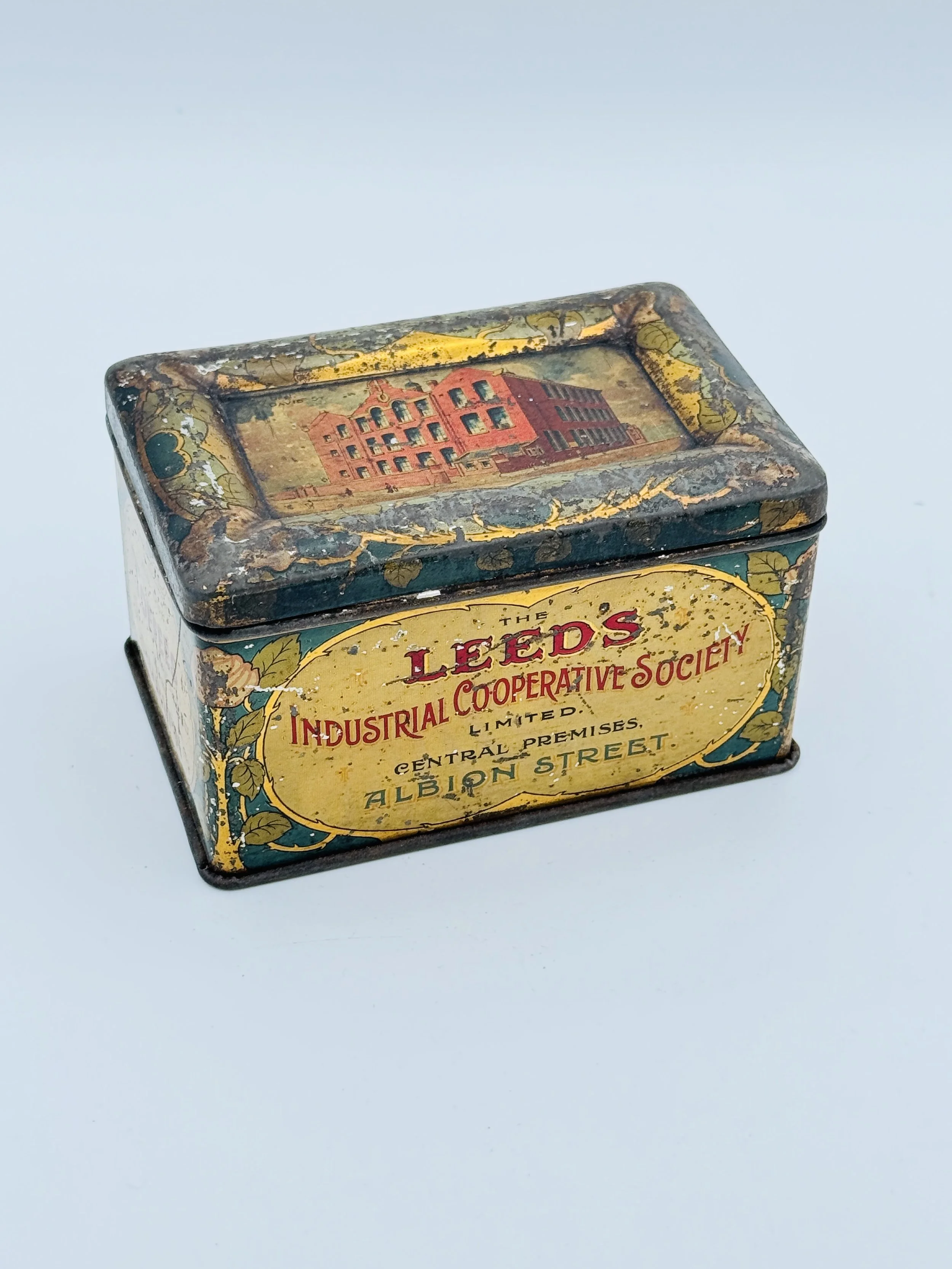 A vintage rectangular metal tin with a worn, decorative design, featuring an illustration of a building on the top and a label on the front that reads 'The Leeds Industrial Cooperative Society Limited, Central Premises, Albion Street.'