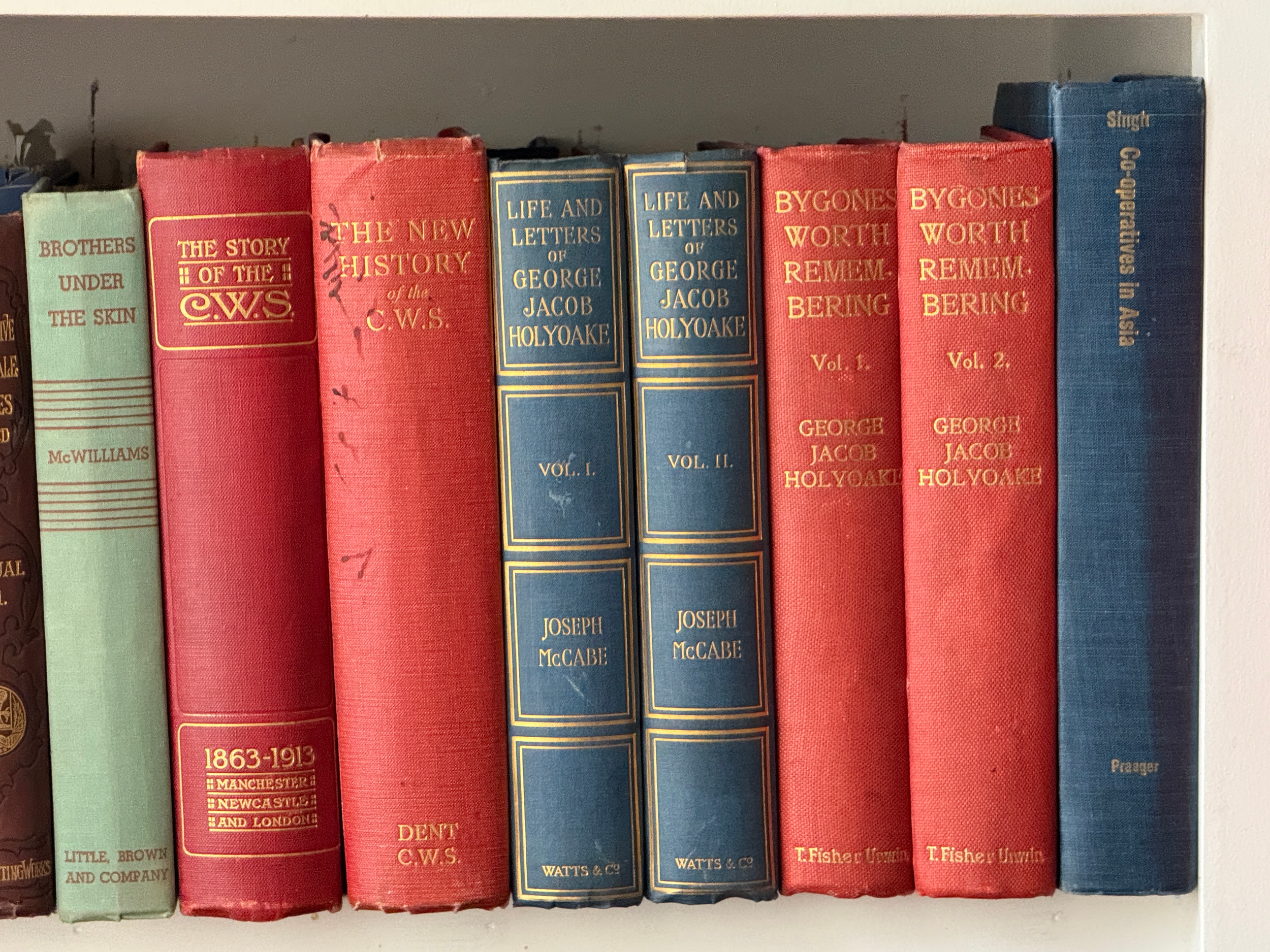 A row of hardcover books on a shelf, with titles including 'Brothers Under the Skin,' 'The Story of the C.W.S.,' 'The New History of the C.W.S.,' 'Life and Letters of George Jacob Holyoake,' and works by George Jacob Holyoake.