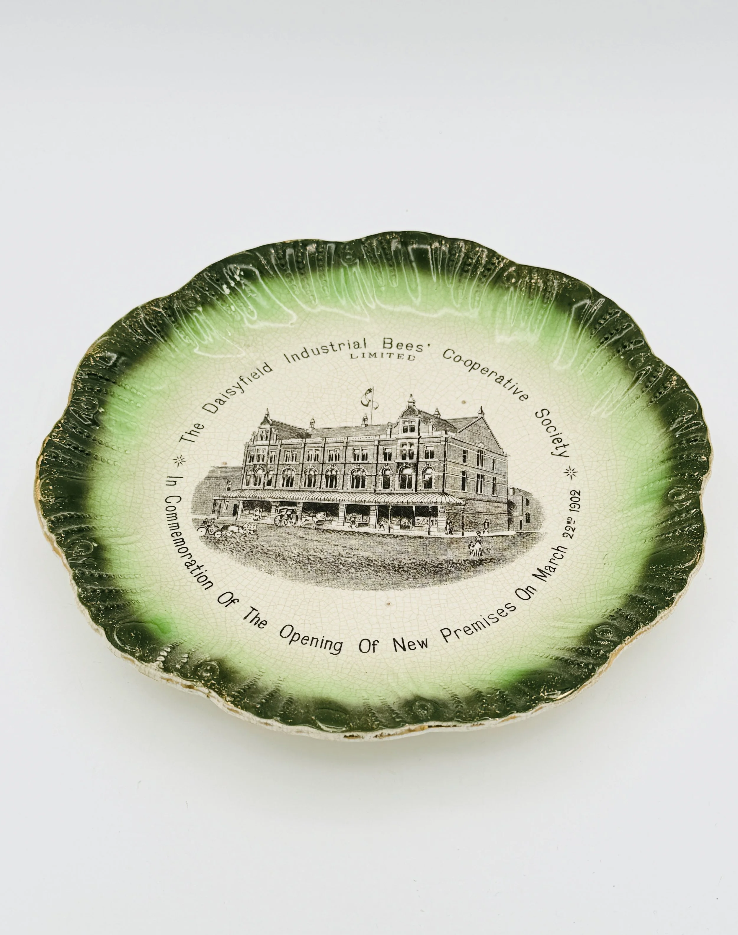 Decorative ceramic plate with green scalloped edge, featuring an old black-and-white illustration of a large building and text about the opening of new premises on March 22, 1902, by the Brisbane Daisyfield Industrial Bees.