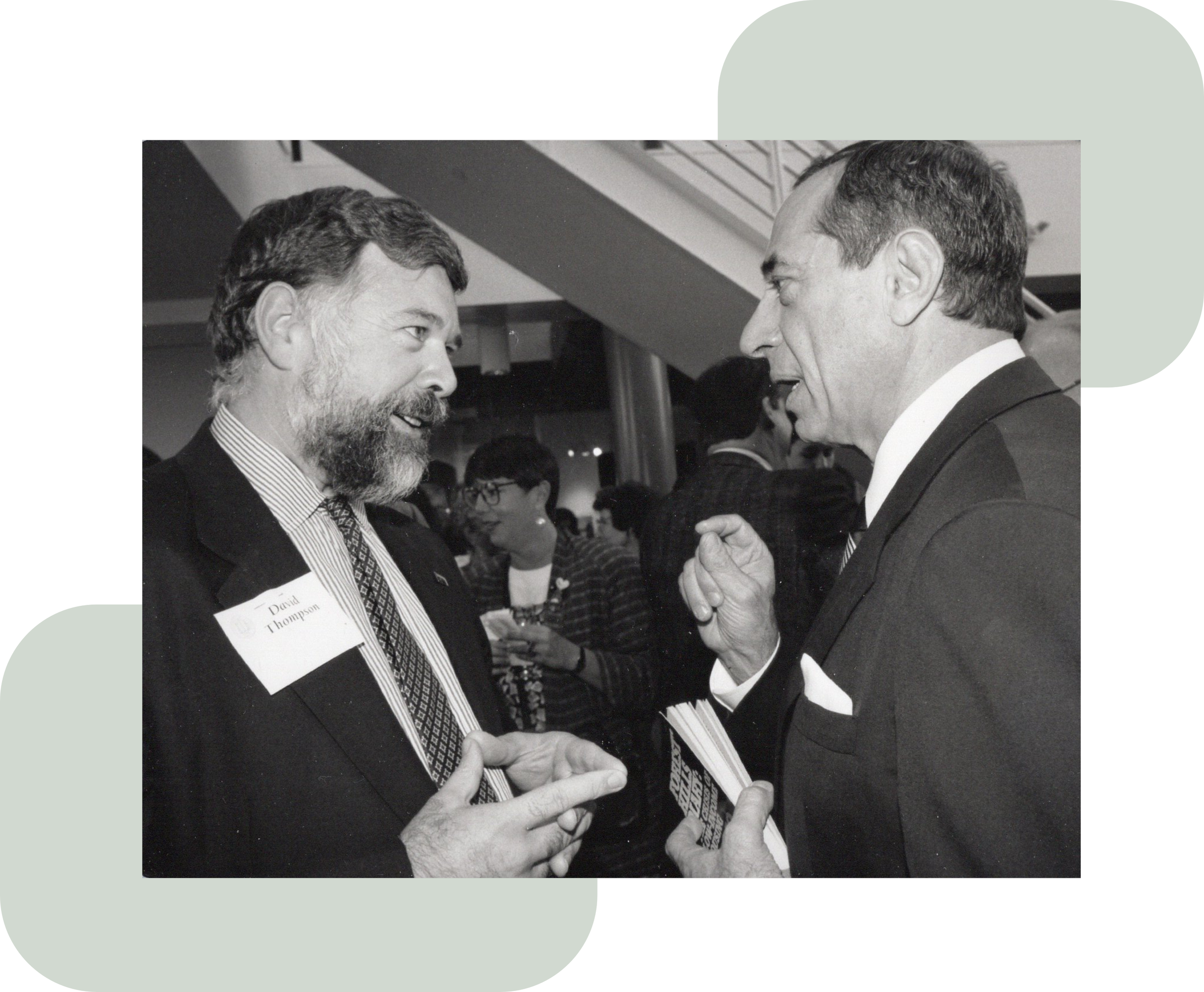 Black-and-white photo of David J Thompson discussing cooperative housing policy with Mario Cuomo at a public social event. Cuomo holds a book in his hand titled "Forest Hills Diary: The crisis of low income housing"