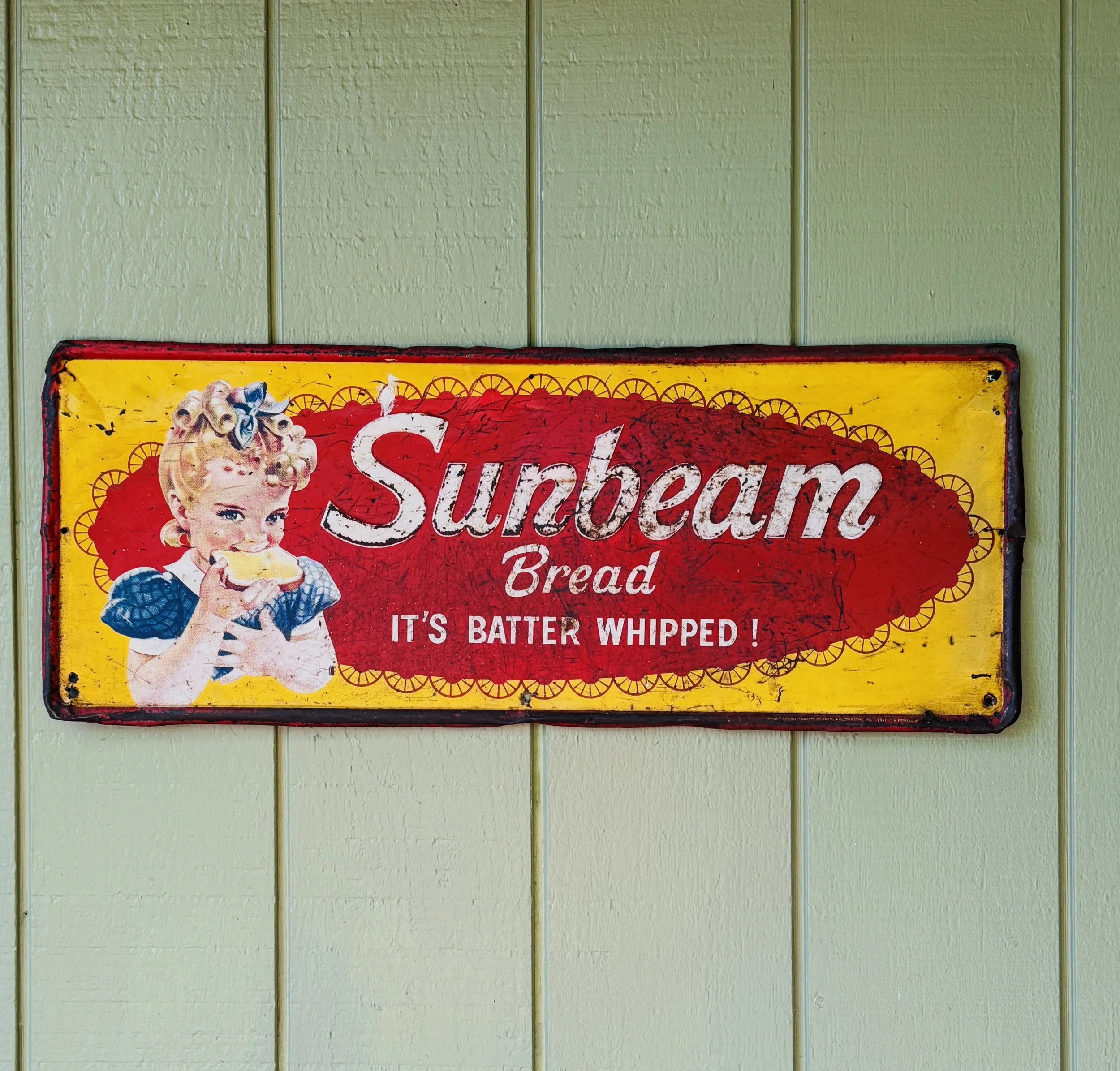 Vintage sign for Sunbeam bread featuring a girl with curlers in her hair eating a toast, with the text 'Sunbeam Bread - It's Batter Whipped!' on a yellow and red background.
