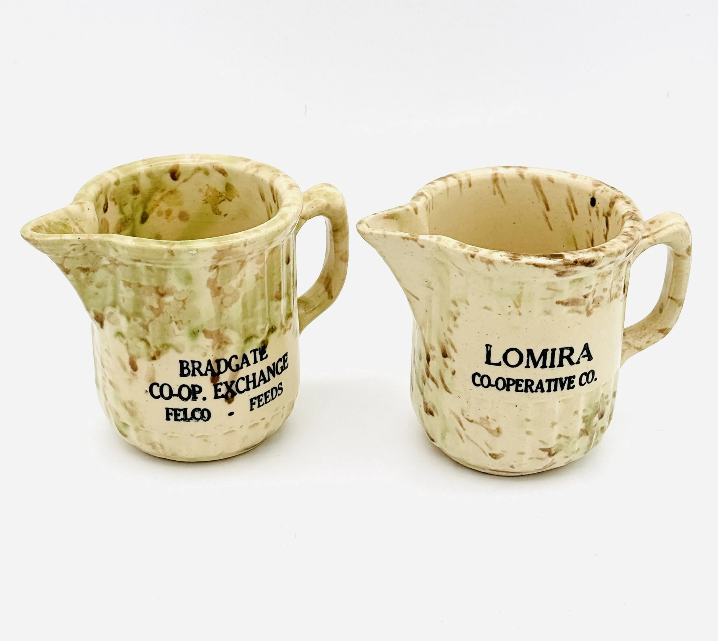 Two small ceramic pitchers with marbled beige and brown patterns, each labeled with black text: one says "BRADGATE CO-OP. EXCHANGE FELCO - FEEDS" and the other says "LOMIRA COOPERATIVE CO.".