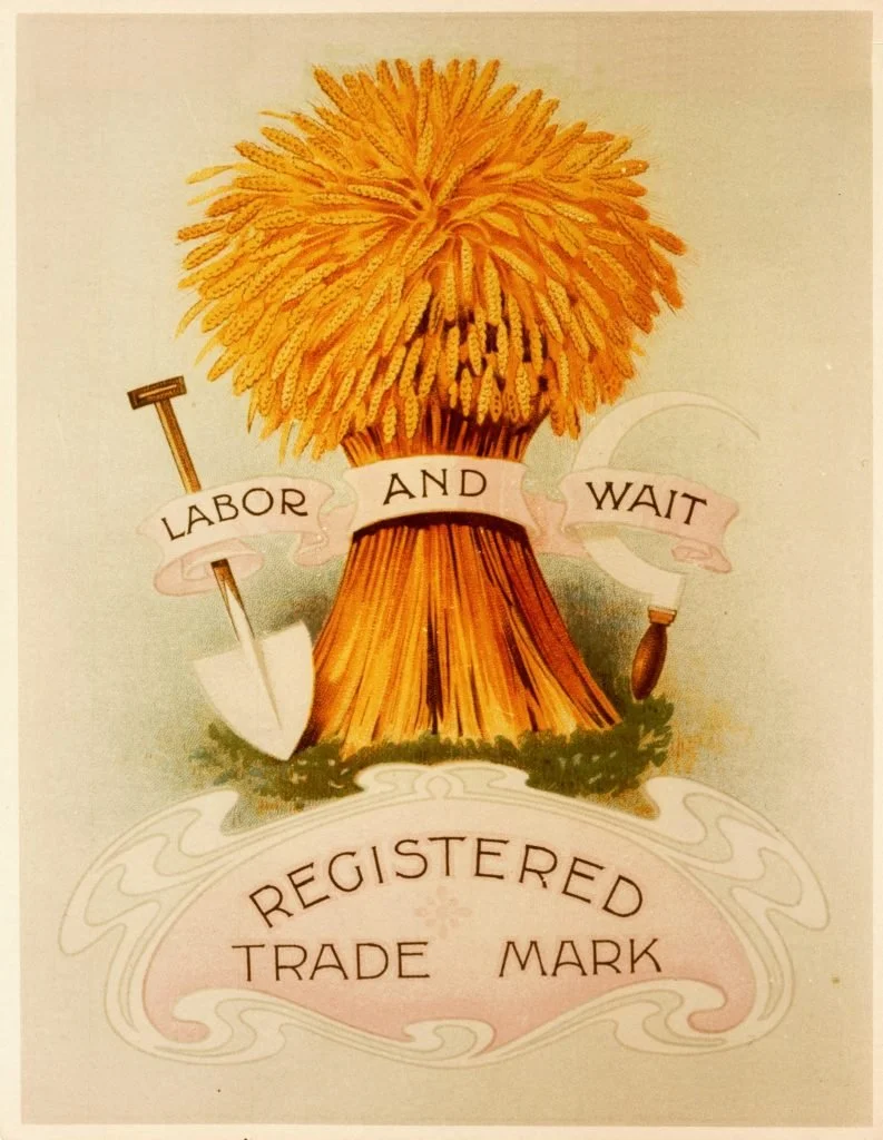 A bundle of wheat tied together with a ribbon that says 'Labor and Wait.' The wheat bundle is adorned with a small shovel on the left and a small axe hanging on the right. The text below reads 'Registered Trade Mark.'
