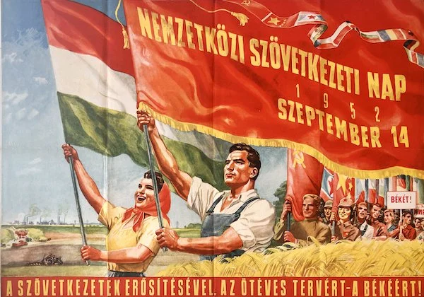 A propaganda poster depicting a man and woman holding a large red flag with yellow text, surrounded by workers and soldiers, promoting International Workers' Day on September 14, 1952, with references to strength through unity and peace.