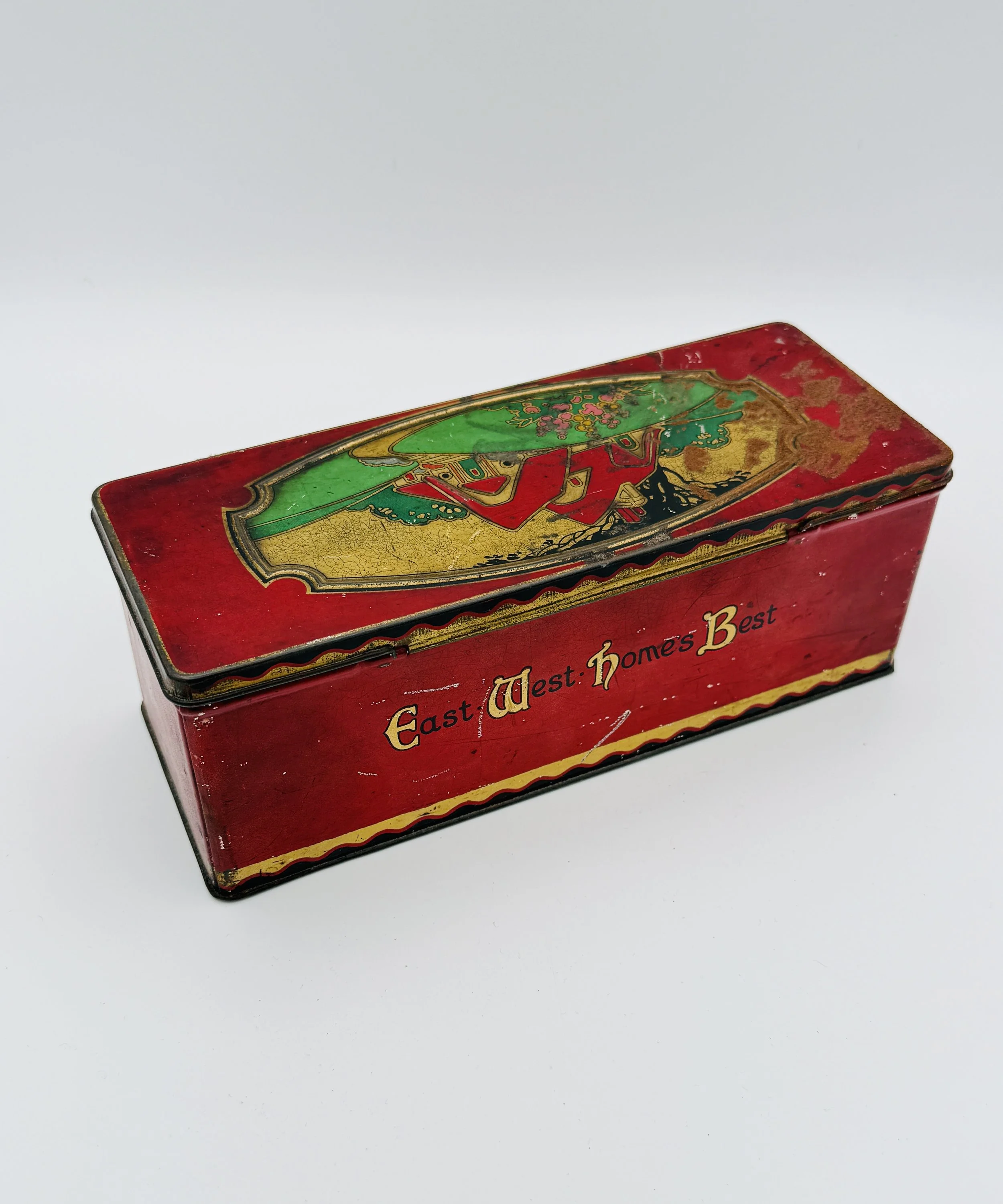 Vintage rectangular red tin box with a colorful image of a woman in traditional dress on the lid, and the text 'East West Home's Best' on the side.