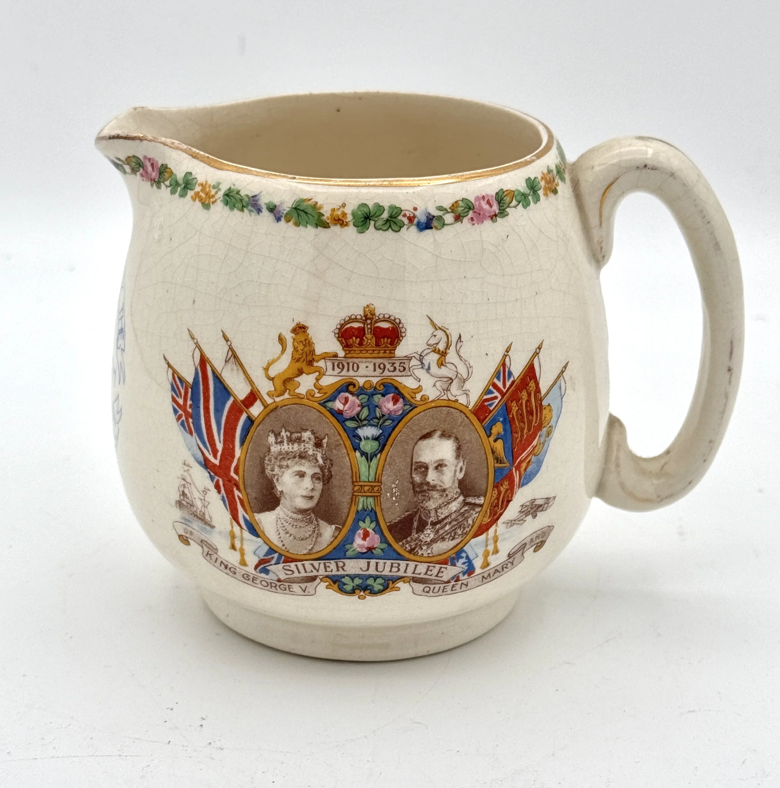 Ceramic mug with a royal commemorative design, featuring portraits of Queen Elizabeth II and King George V, surrounded by flags, floral decorations, and the text for the Silver Jubilee 1910-1935.