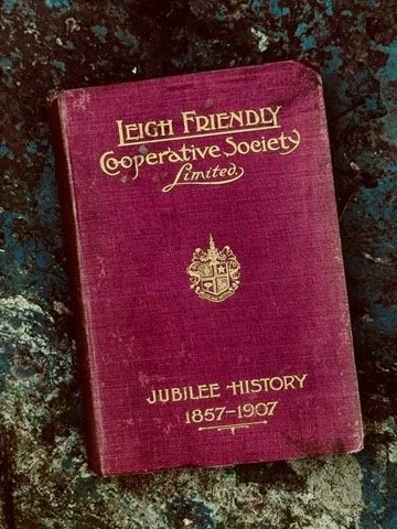 A maroon hardcover book titled 'Leigh Friendly Cooperative Society Limited Jubilee History 1857-1957' with a gold emblem on the cover, resting on a dark, textured surface.