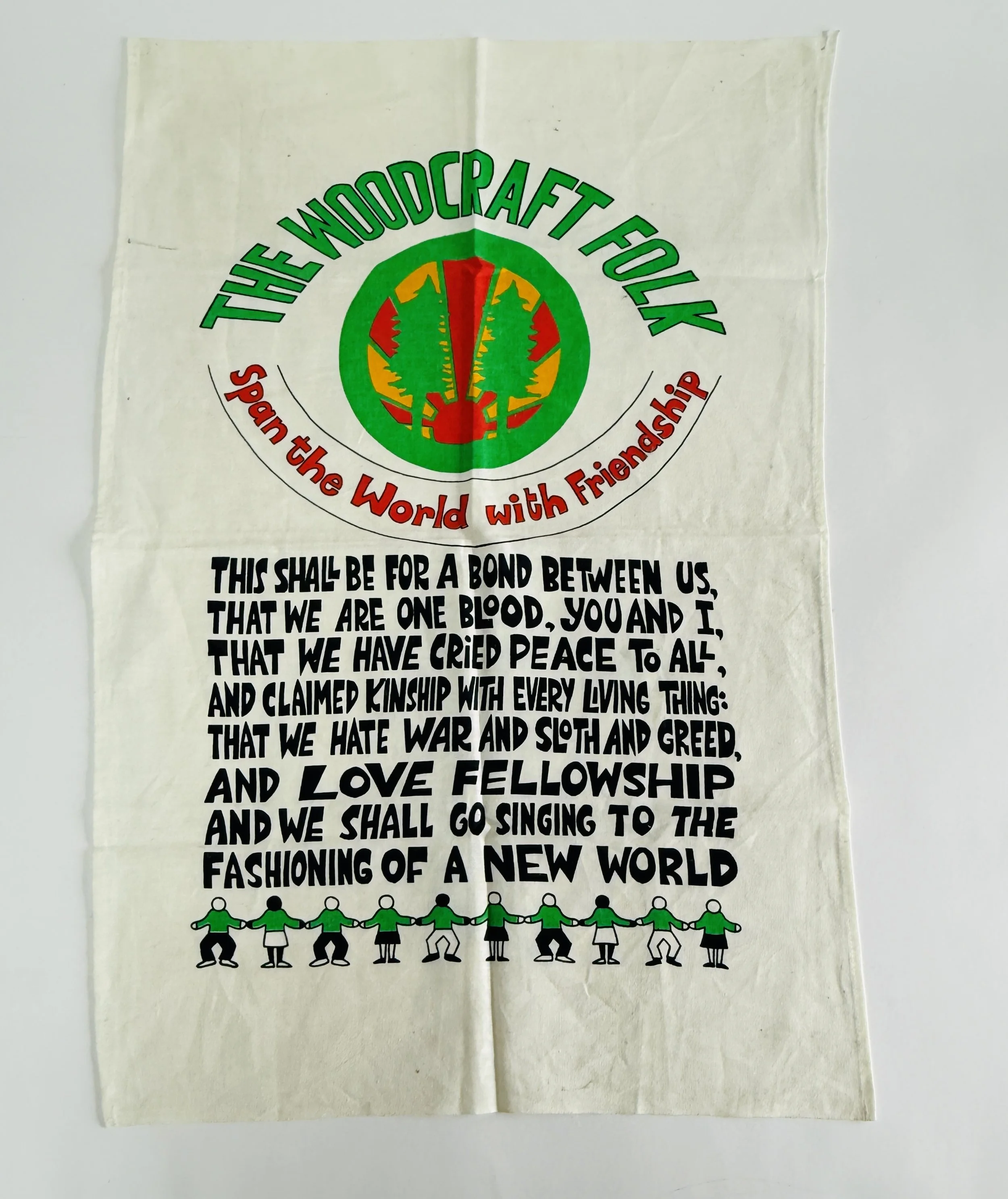 A cloth banner with the words "The Woodcraft Folk" at the top, a logo with trees and a red and green circle, and the slogan "Span the World with Friendship." Below, a poetic excerpt about peace, love, and unity, with a row of cartoon children holding