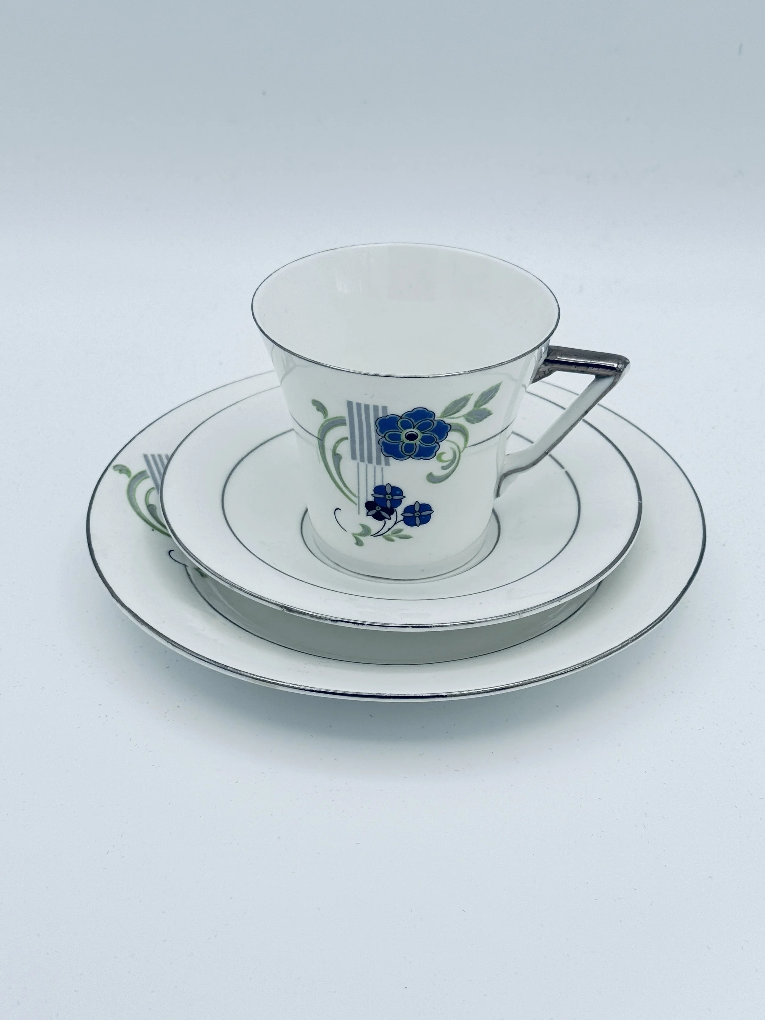 A set of three white porcelain dishes with blue and green floral patterns, including a teacup with a handle, a small plate, and a larger plate, all stacked on a white surface.