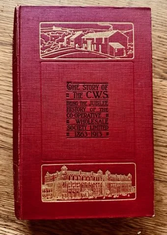 A red hardcover book with gold embossed illustrations of an industrial scene and a building on the cover, and black text reading 'The story of the C&W's Being the Julie's History of the Co-Operative & Wholesale Society Limited 1863-1933'.