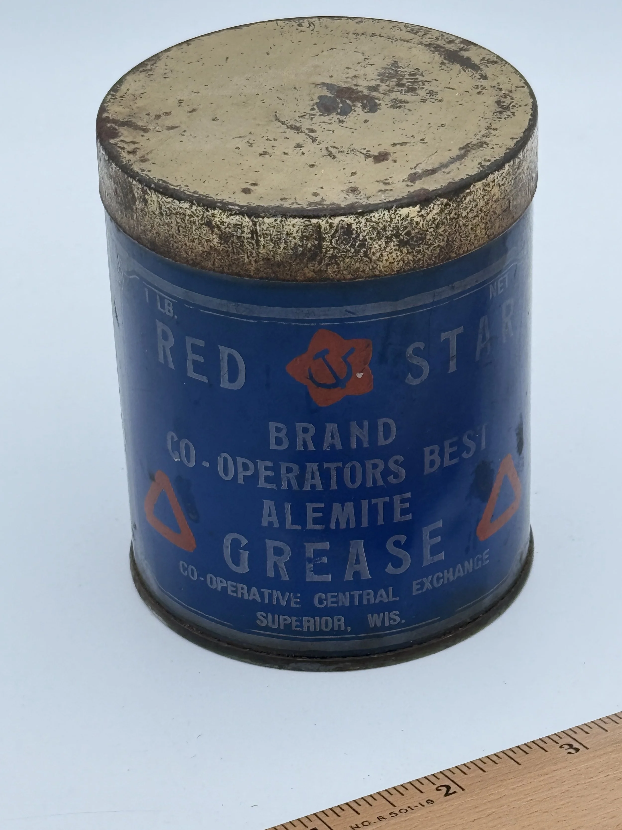 A vintage blue tin container labeled 'Red Star Brand Co-Operators Best Alemite Grease' with a red star and blue anchor logo, showing signs of rust and age, placed on a white surface with a ruler nearby for scale.