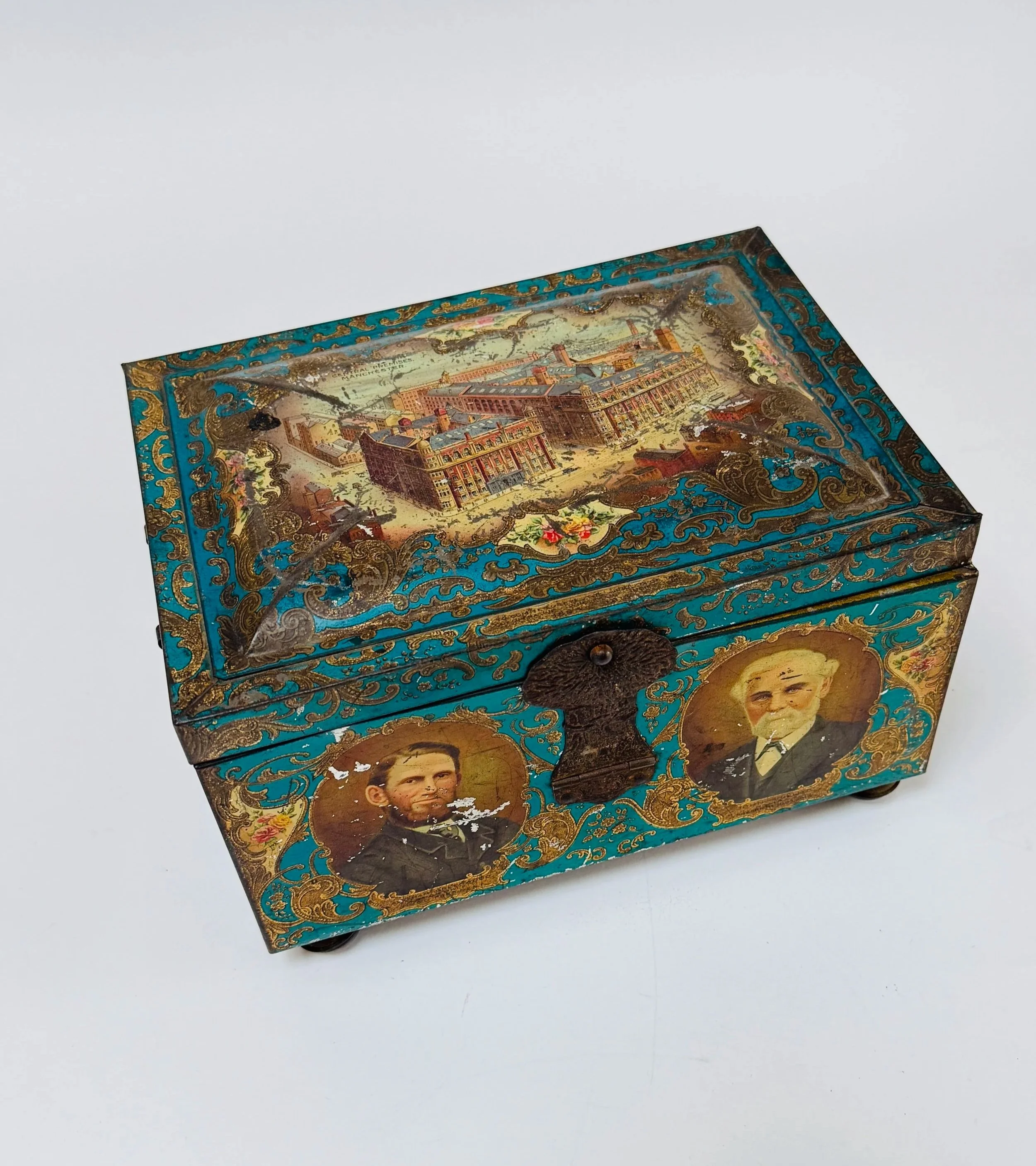 Decorative antique tin box with painted portraits of two men and a detailed scene of a large building complex on the lid.