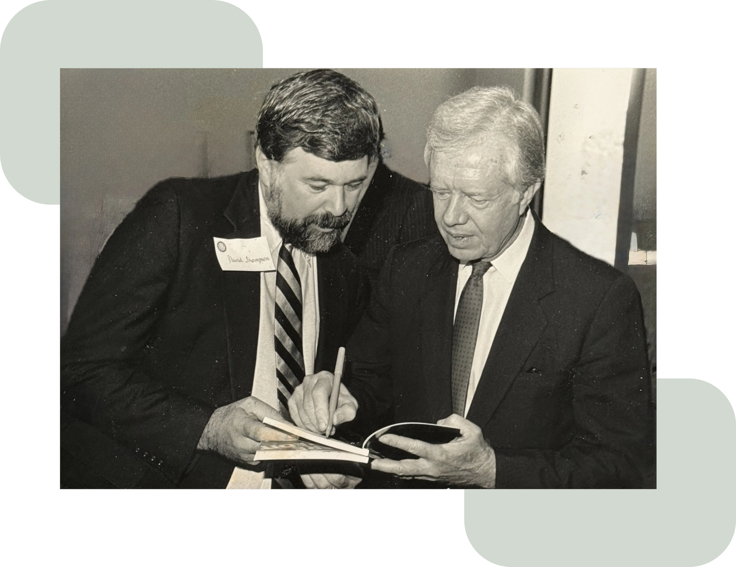 Black-and-white photo of two men in suits reading and signing a book; the younger man standing on the left wears a name tag reading “David Thompson.”