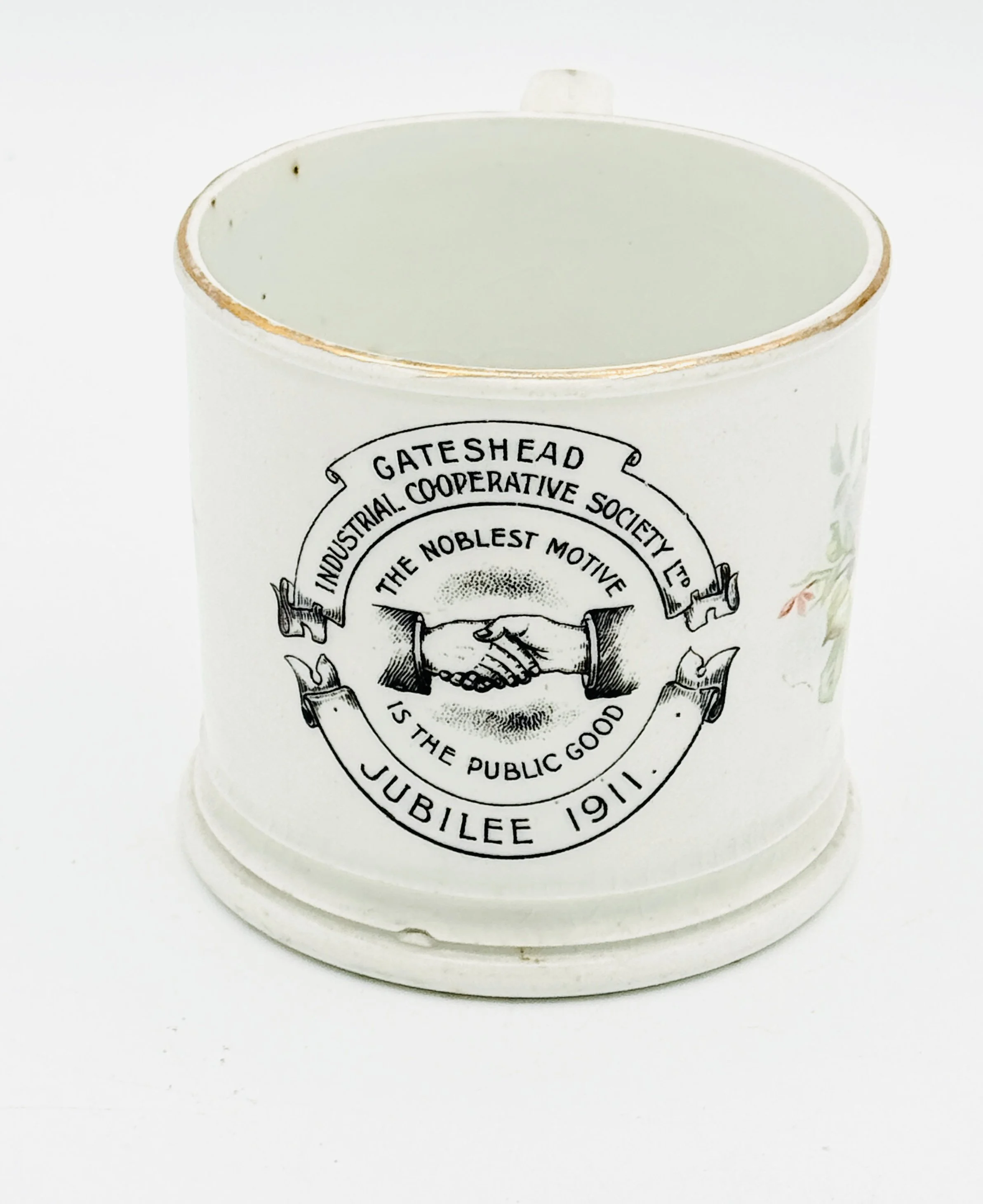 A white ceramic mug with a vintage black and white logo of the Gateshead Industrial Cooperative Society, featuring a handshake, the text 'The Noblest Motive is the Public Good,' and details about a Jubilee in 1911, placed on a white background.